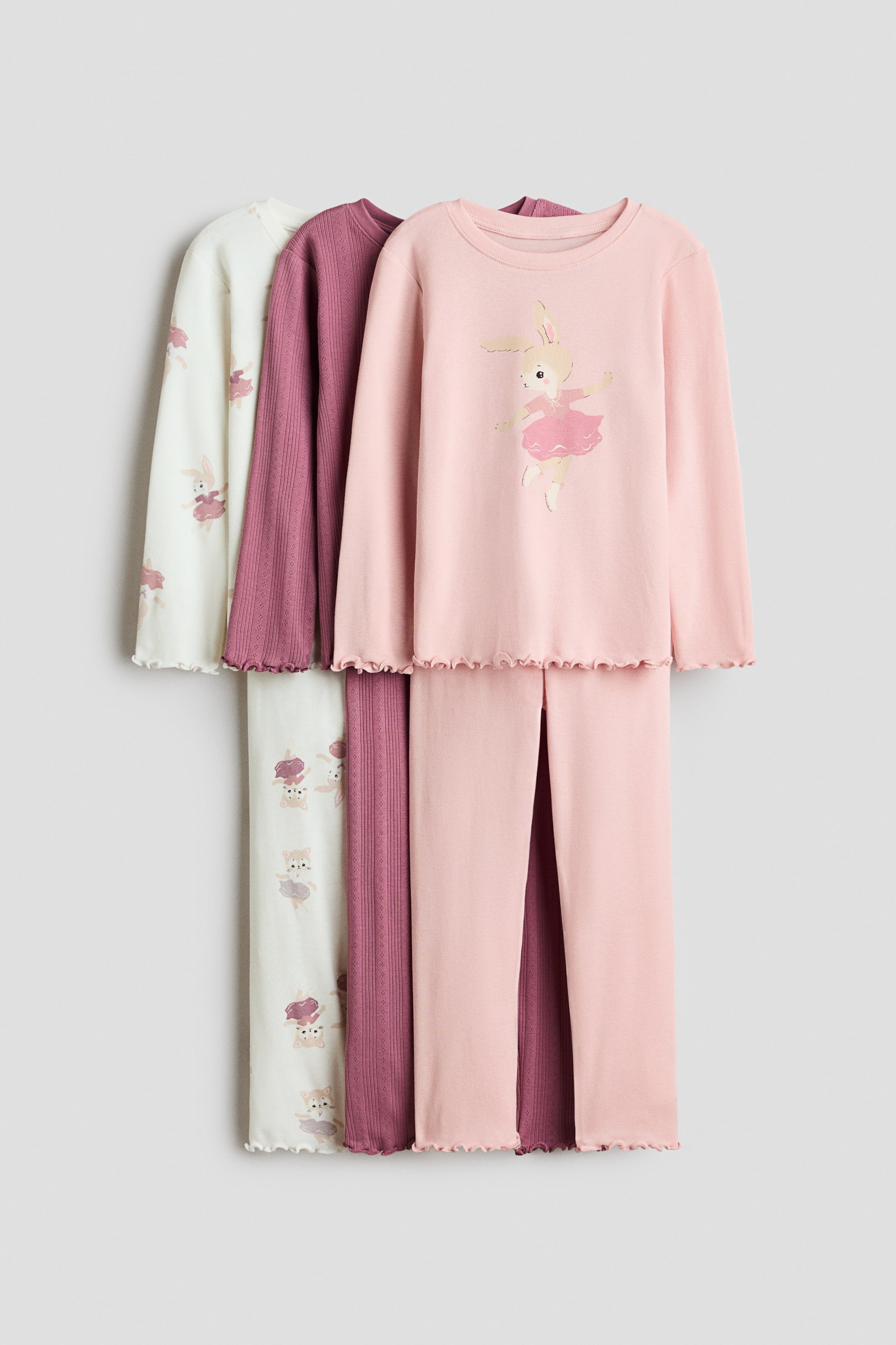 Größeres Bild anzeigen: Drei Jersey-Pyjama-Sets aus Baumwolle hängen gemeinsam nach vorne ausgerichtet und bestehen aus Langarmoberteilen und passenden Hosen mit rollierten Lettuce-Säumen. Das vordere Set ist hellrosa mit einem Ballerina-Häschen-Motiv, das mittlere Set ist einfarbig dunkelrosa in Rippstrick, und das hintere Set ist cremeweiß mit einem allover Hasen-Print.