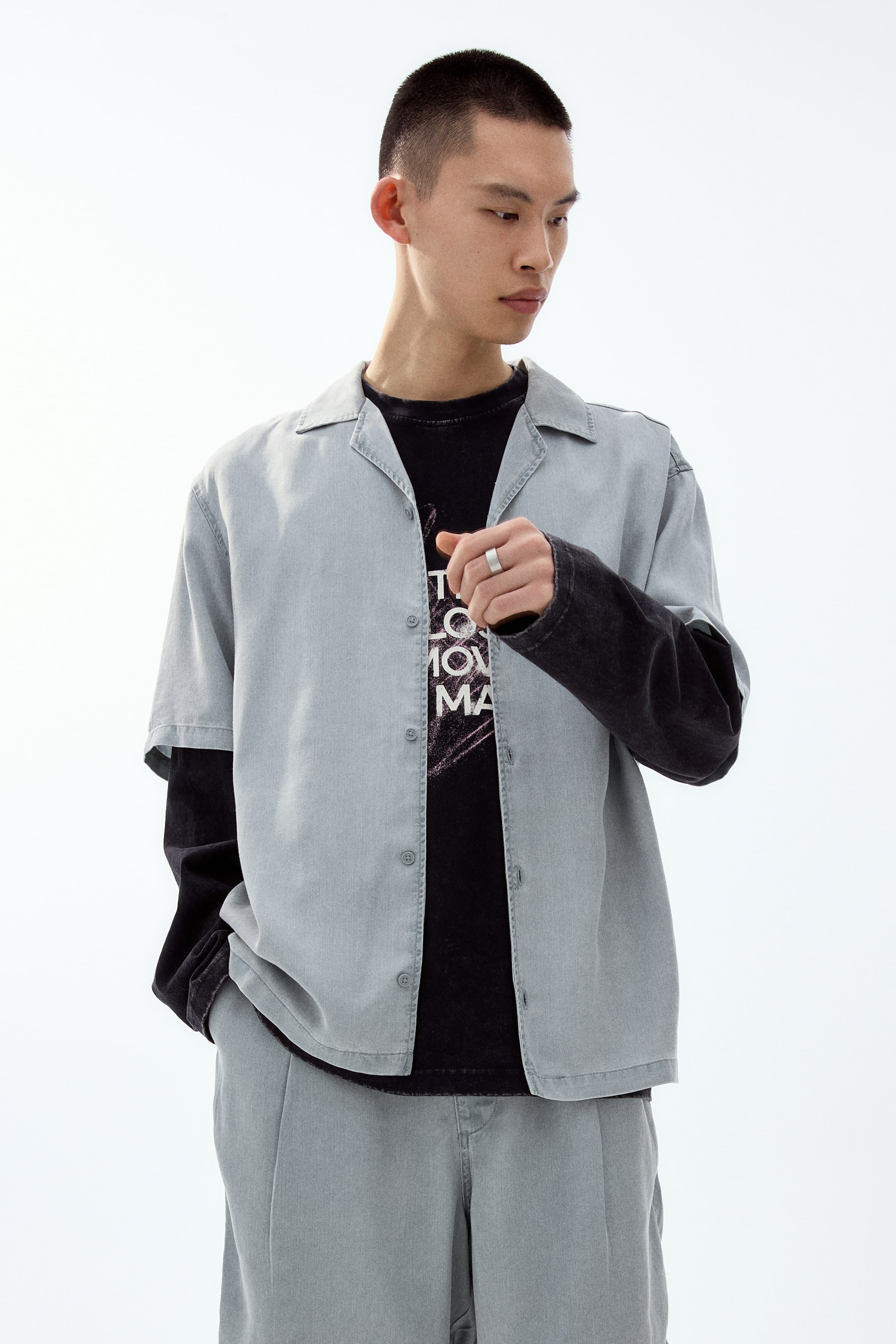 View larger image: A light grey short-sleeved shirt features a camp collar and wide short sleeves. It has an open button-front placket and a straight hem, with a relaxed fit that drapes over a black long-sleeved shirt.