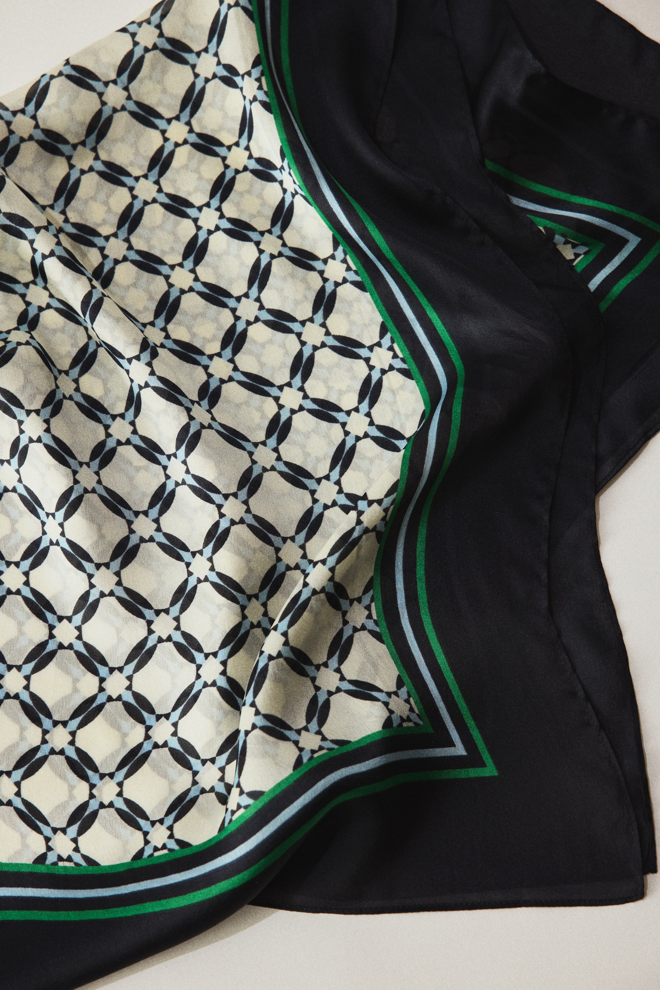 View larger image: The scarf features a geometric pattern of black interlocking shapes with light-blue accents on a cream background, bordered by a wide black band with thin green and light-blue stripes.
