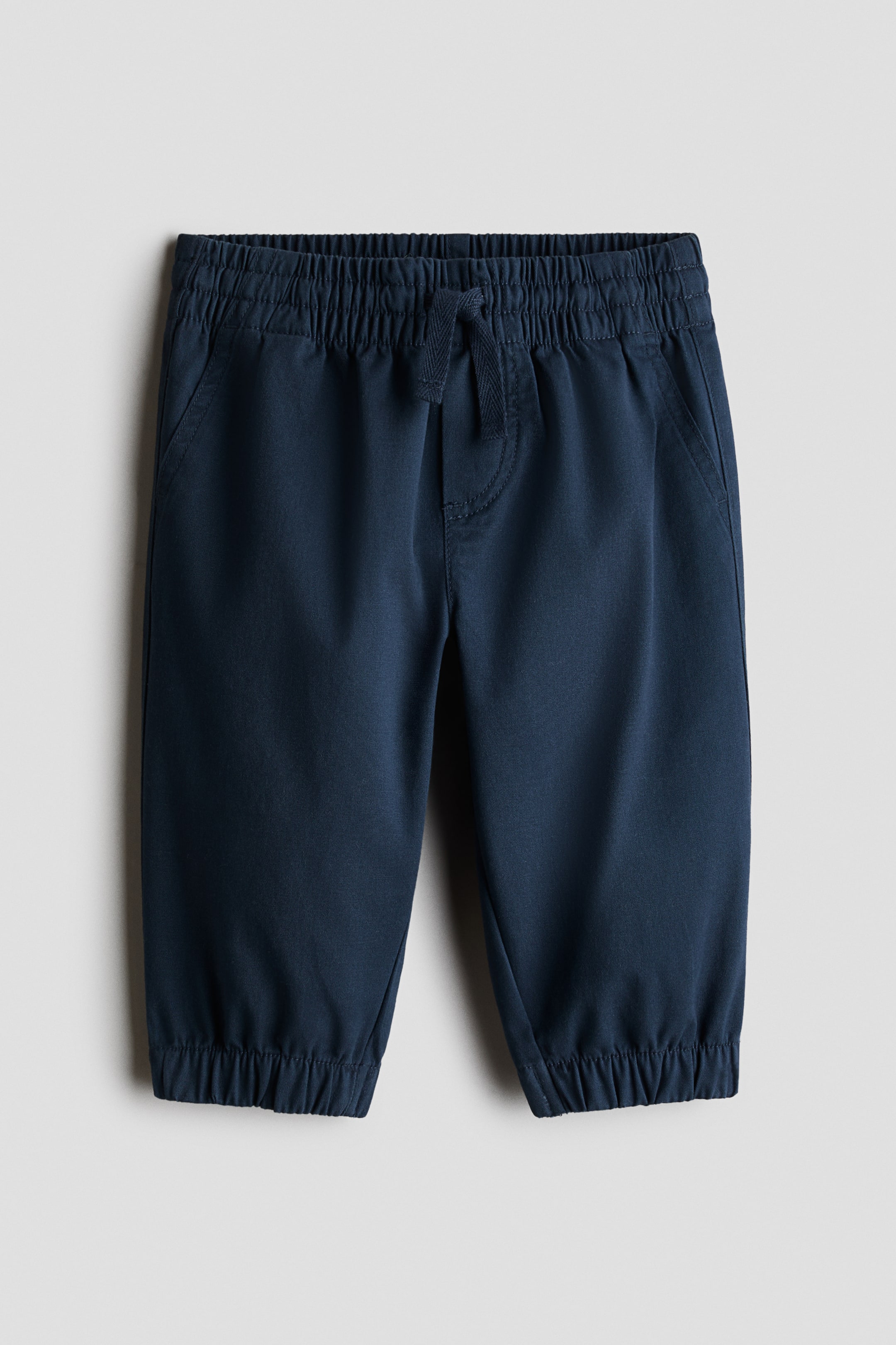 View larger image: Presenting a pair of navy blue cotton trousers from a 3-piece ensemble, showcased from the front. These trousers feature an elasticated waistband with a visible drawstring tie, side pockets, and elasticated jogger cuffs.
