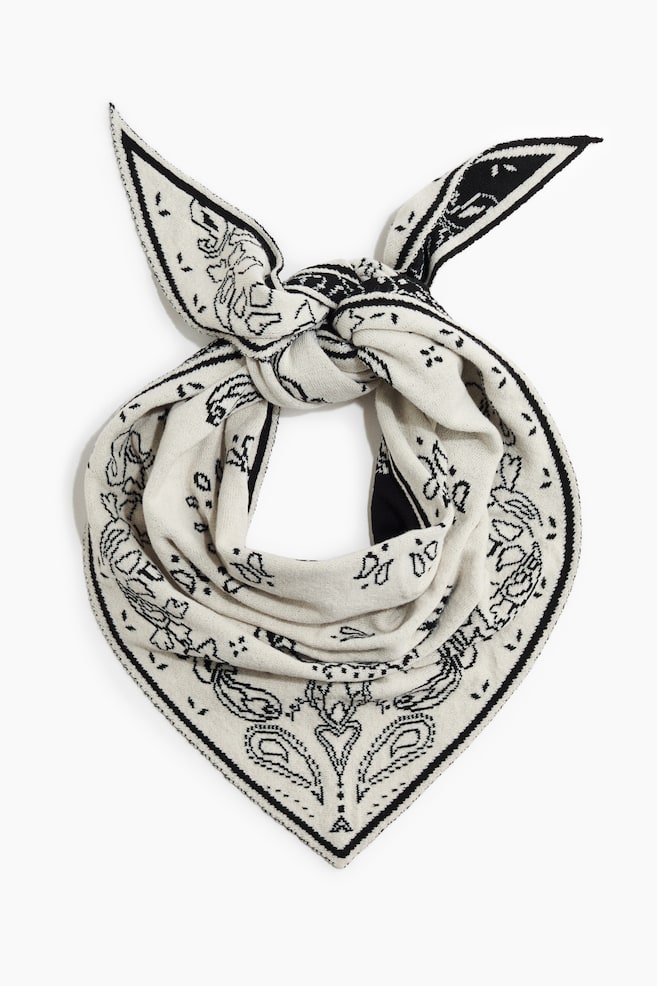 Ein cremefarbener Strick-Bandana-Schal mit schwarzem Paisley-Muster ist aus der Vorderansicht zu sehen. Der Schal ist oben zu einem Knoten gebunden, hat zwei spitze Enden und fÃ¤llt in einer dreieckigen Form nach unten. - 1