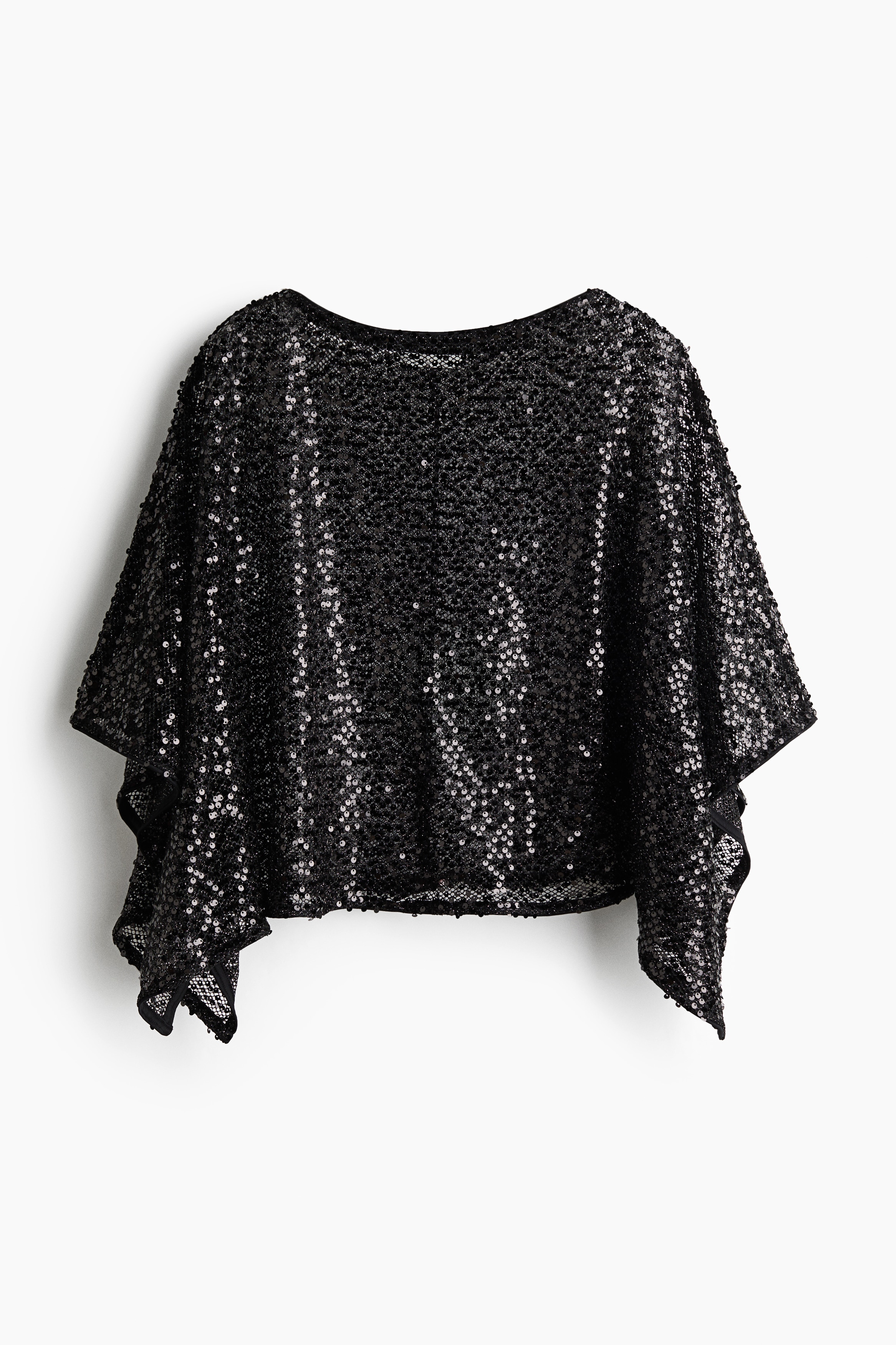 An oversized cape top in a sheer, sequin-covered knit is worn by the model over dark undergarments and trousers. The top features a delicate boat neck and a draped handkerchief hem, highlighted as the model stands looking down with one hand resting near her hip.