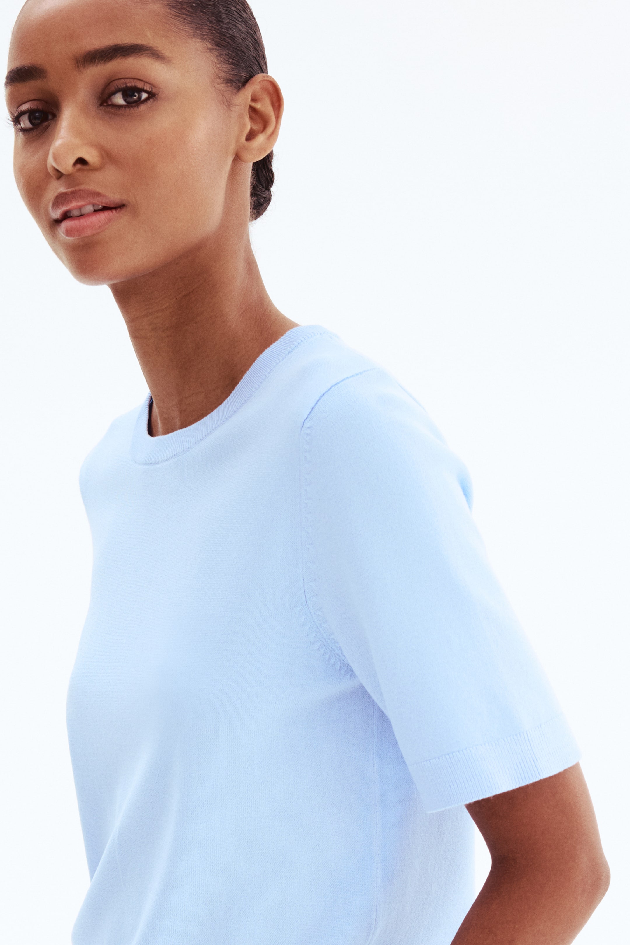 Xem ảnh lớn hơn: Người mẫu đang mặc một áo dệt kim mềm màu xanh nhạt, có cổ tròn, tay ngắn và viền rib ở cổ và ống tay.