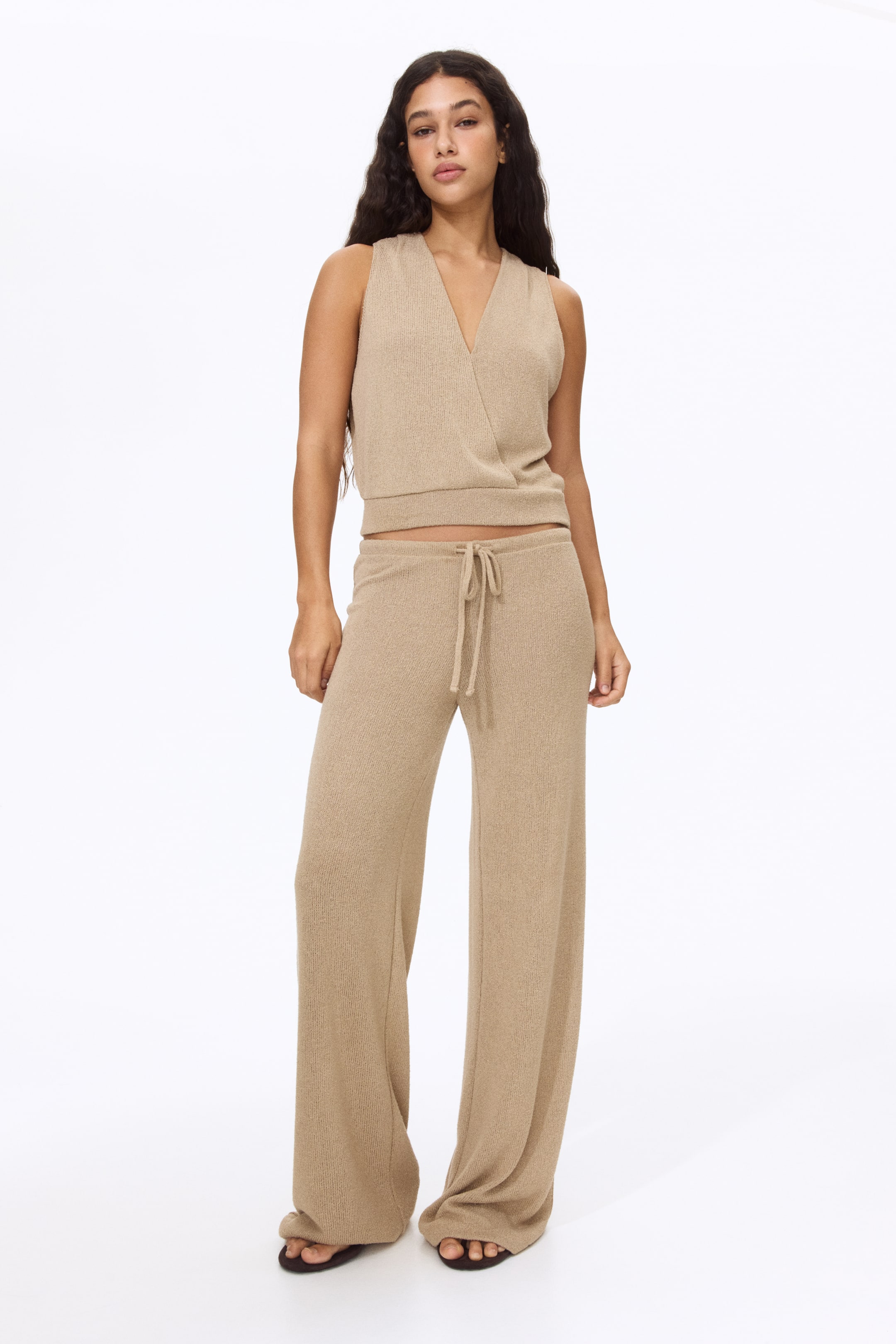 View larger image: Presenting a beige knitted two-piece ensemble, displayed facing forward. The sleeveless top features a V-neckline, a wrap-style front, and a ribbed band at the hem, while the wide-leg trousers boast a drawstring waist and a matching knitted texture.