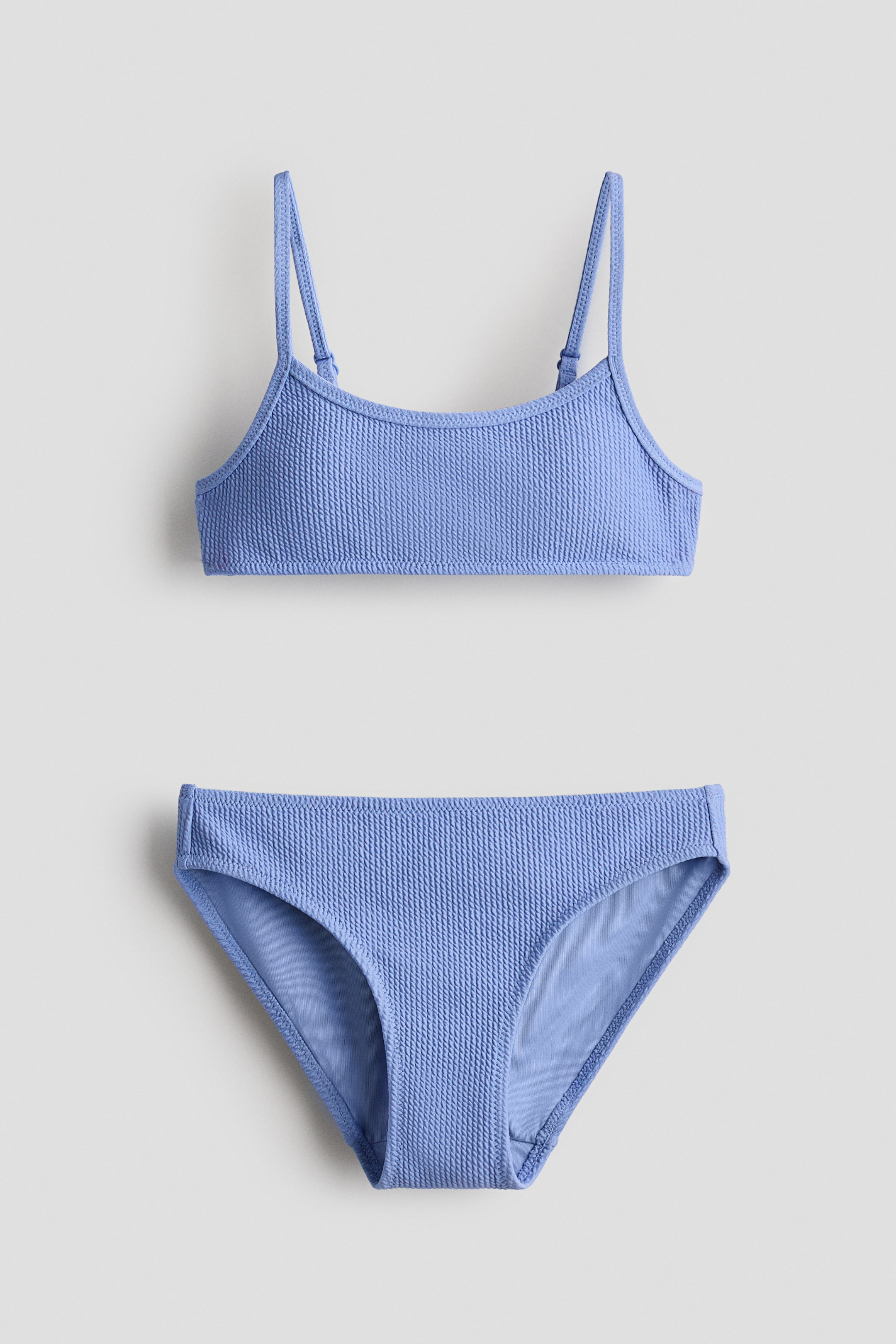 Grotere afbeelding bekijken: Een periwinkle blauwe tweedelige badpak met een gestructureerde kreukelstof. De bralette-stijl top heeft dunne verstelbare spaghetti bandjes en een ronde halslijn, en de bijpassende klassieke slip heeft een standaard snit.