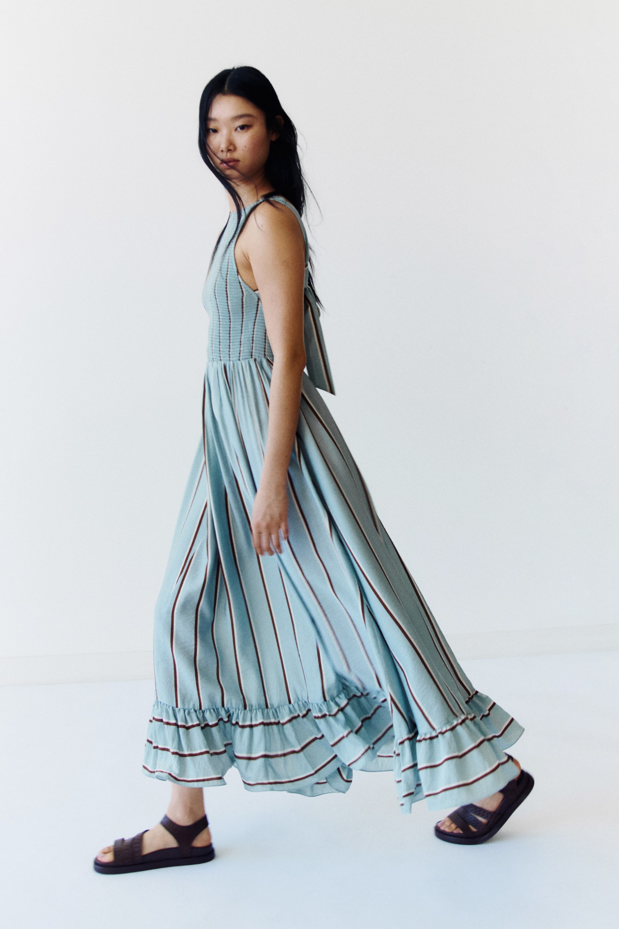 View larger image: The light blue maxi dress, viewed from a side-front angle, features thin brown and white vertical stripes. It is crafted with a smocked bodice, slender shoulder straps that tie into a bow at the back, and a full, flowing skirt ending in a ruffled hem.