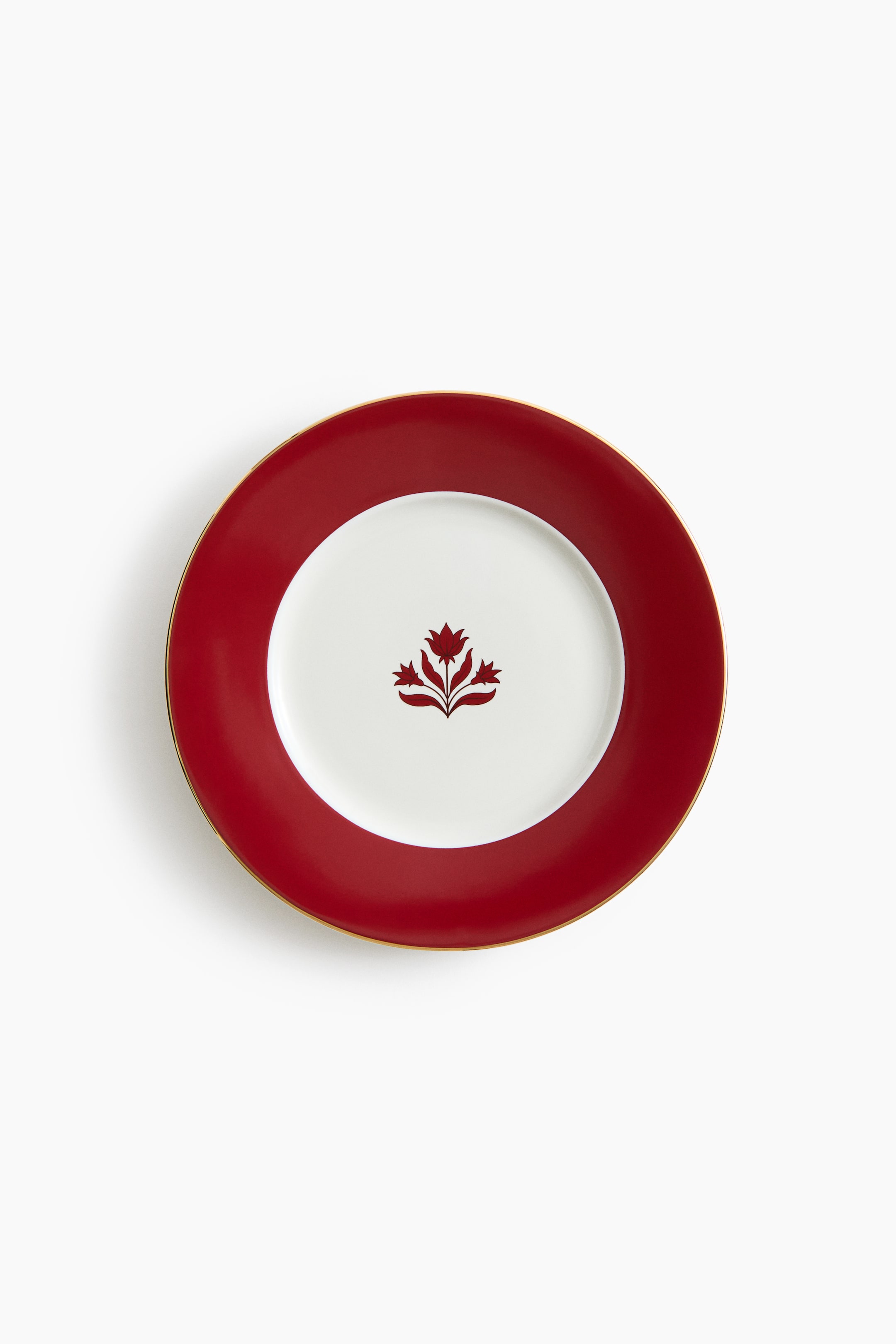 View larger image: A round ceramic plate is shown from a top-down angle, featuring a white centre decorated with a stylised red floral motif and surrounded by a wide red border with a thin gold rim.