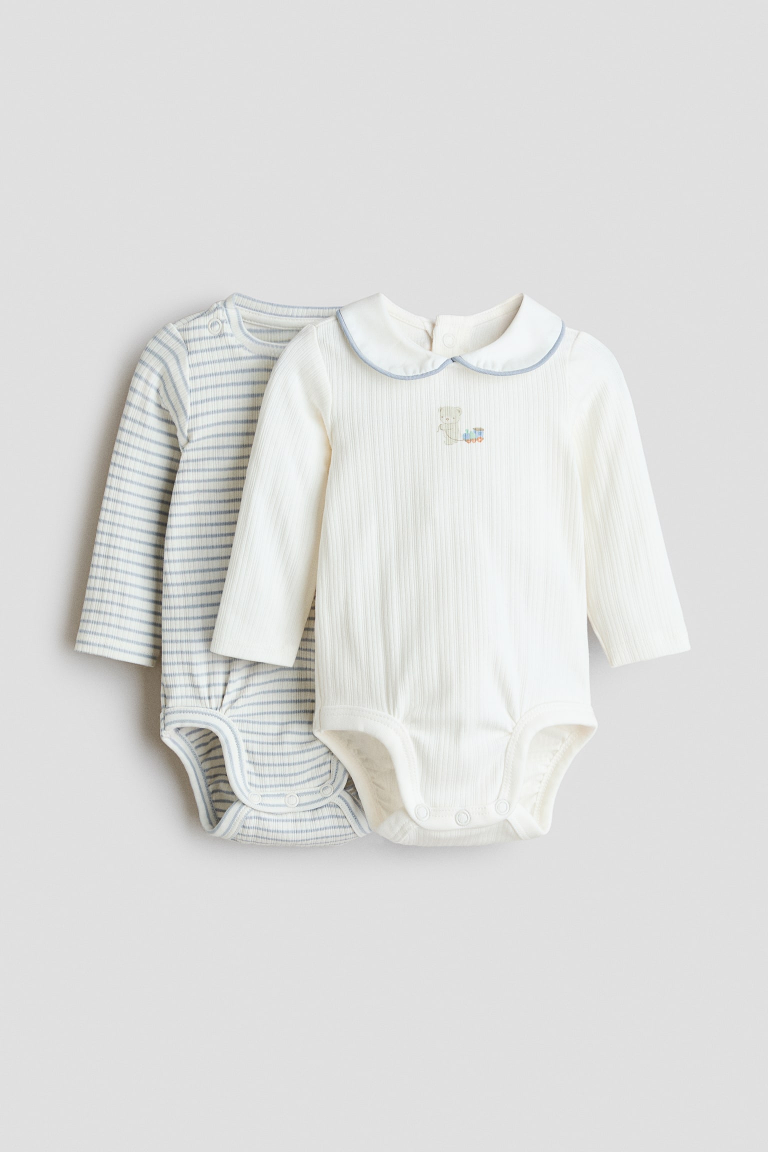 Zwei langÃ¤rmelige Baby-Bodysuits werden von vorne gezeigt und verfÃ¼gen Ã¼ber gerippte Strickstoffe sowie DruckknopfverschlÃ¼sse im Schritt. Der vordere Bodysuit in Off-White ist mit einem Peter-Pan-Kragen mit hellblauer Paspelierung und einem kleinen bestickten Motiv auf der Brust versehen, wÃ¤hrend der dahinter liegende Bodysuit ein hellblau-weiÃes horizontales Streifenmuster aufweist.