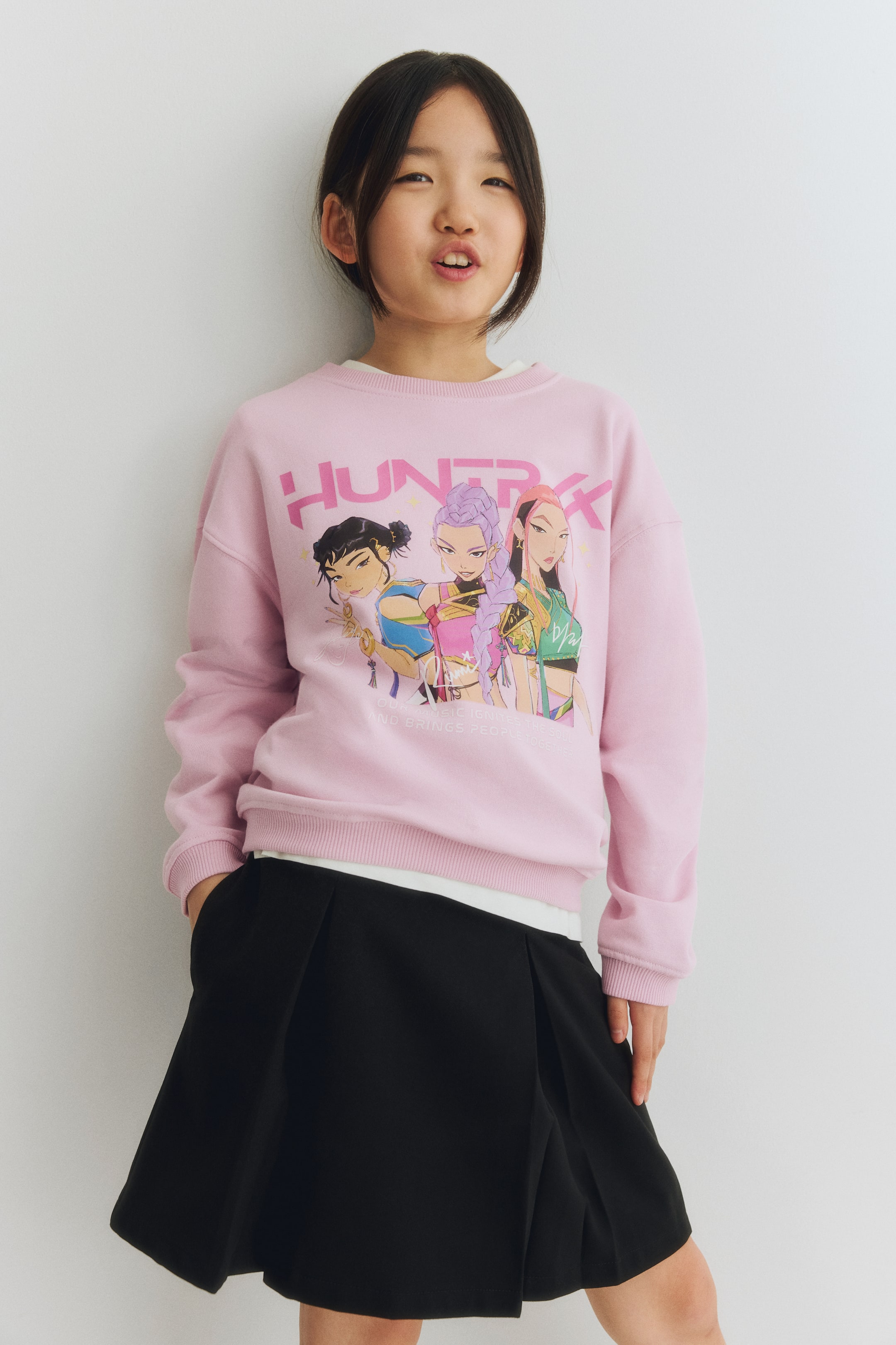 View larger image: A pink crew-neck sweatshirt featuring a large graphic print on the front, displaying "HUNTRXX" in pink letters, three animated female characters, and the text "OUR MUSIC IGNITES THE SOUL AND BRINGS PEOPLE TOGETHER". Its ribbed cuffs and hem are visible, and it is worn over a white T-shirt.
