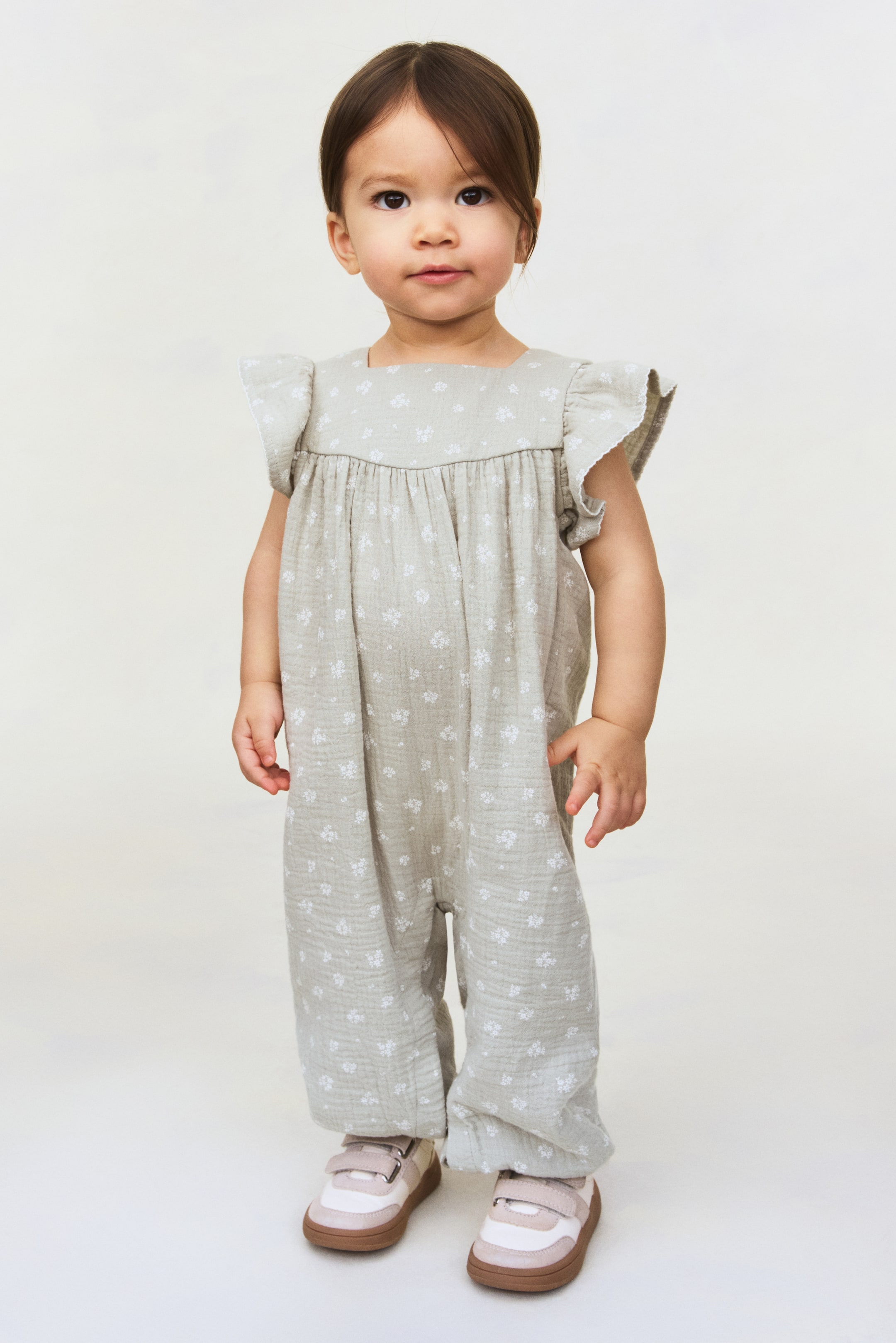 Ver imagen más grande: Un mono verde claro para niño pequeño presenta un pequeño estampado floral blanco, un escote cuadrado y manguitas con volantes. Incorpora un detalle fruncido en el pecho y perneras largas y holgadas, y se presenta combinado con zapatillas rosas, blancas y beige con cierre de velcro.