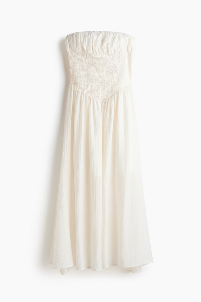 Ein cremefarbenes, trägerloses Maxikleid ist von vorne sichtbar und zeichnet sich durch ein gesmoktes Oberteil aus, das einen elastischen, texturierten Effekt erzeugt und eine V-Form an der Taille bildet. Der lange, fließende Rock besteht aus einem leichten Stoff mit dezenten vertikalen Knitterfalten. - 1