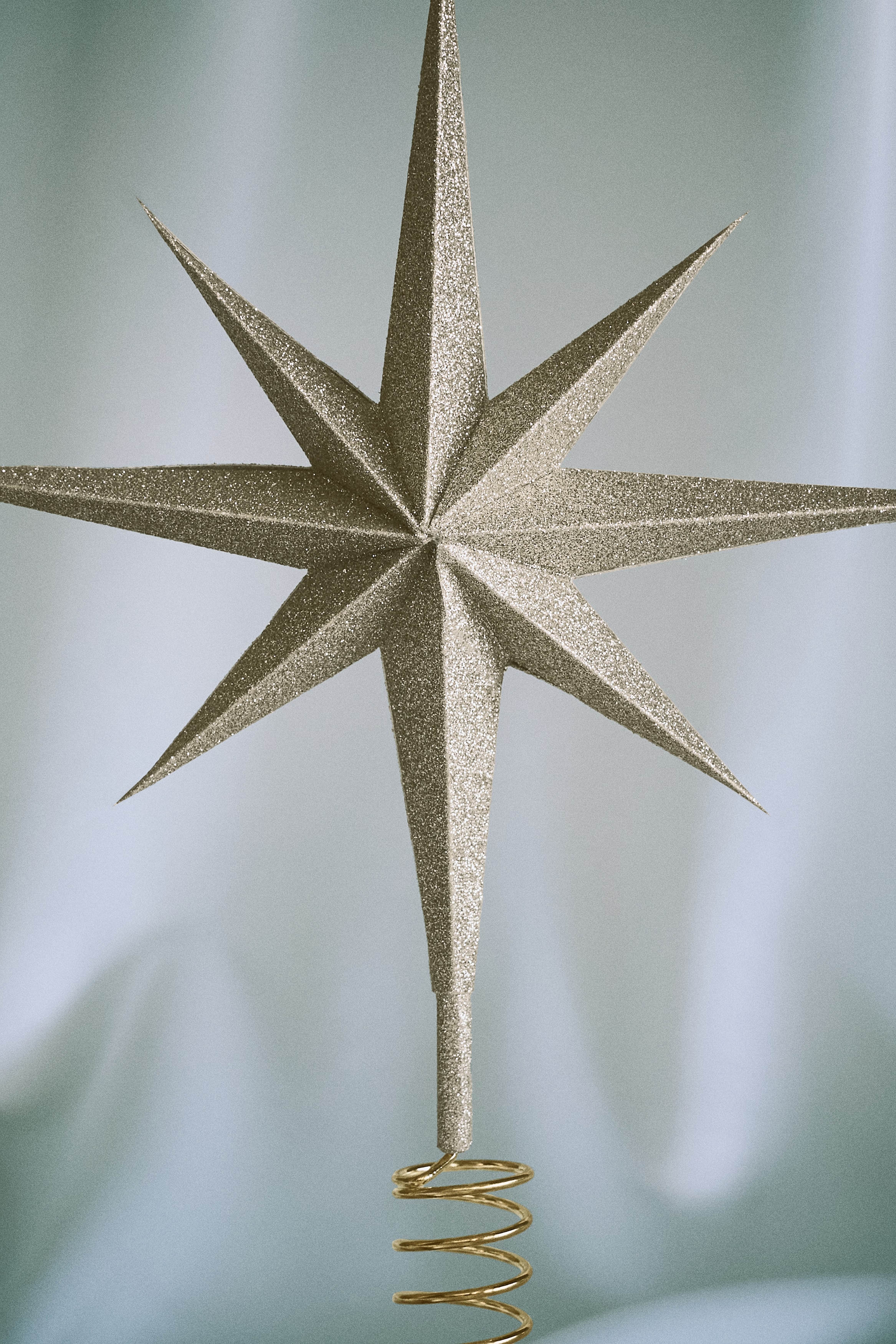 A gold-glittered paper star tree topper is facing front, showcasing an eight-pointed, three-dimensional faceted structure. The bottom point of the star is extending down to connect with a gold-toned metal spiral wire coil base.