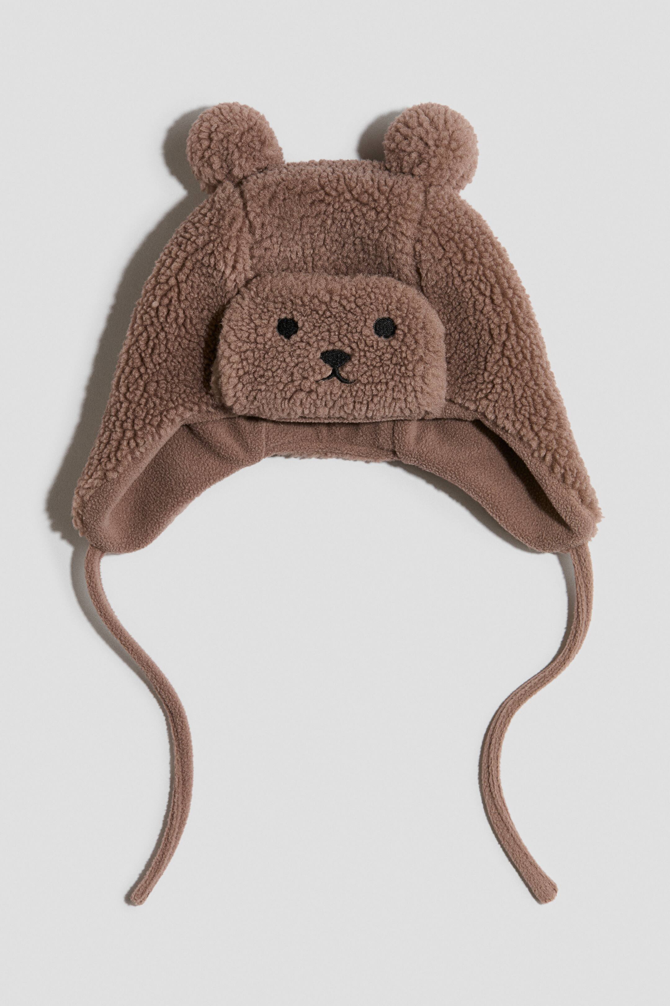 Größeres Bild ansehen: Eine braune Teddybärenmütze präsentiert eine plüschige, sherpaähnliche Textur mit zwei abgerundeten Ohren oben. Sie verfügt über einen erhöhten Frontaufnäher mit einem aufgestickten schwarzen Bärengesicht, während zwei Bänder von den Ohrklappen herabhängen.