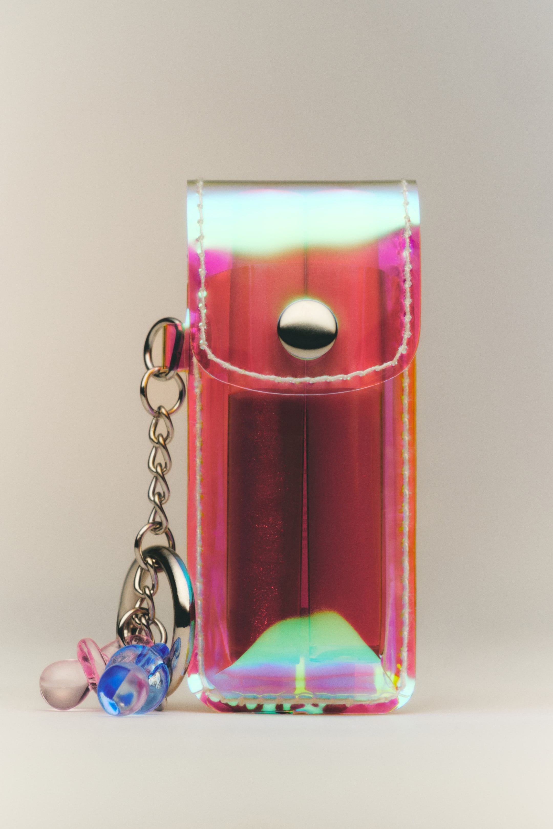 Grotere afbeelding bekijken: Een sleutelring toont een holografisch etui dat rechtop staat en naar voren is gericht. Het etui, met iriserende roze, blauwe en groene tinten, is omzoomd met witte stiksels, wordt gesloten met een zilverkleurige drukknoop en bevat duidelijk twee mini-lipglosses in verschillende tinten. Twee doorschijnende dummy-bedels, één roze en één blauw, hangen aan de ketting van de sleutelring.
