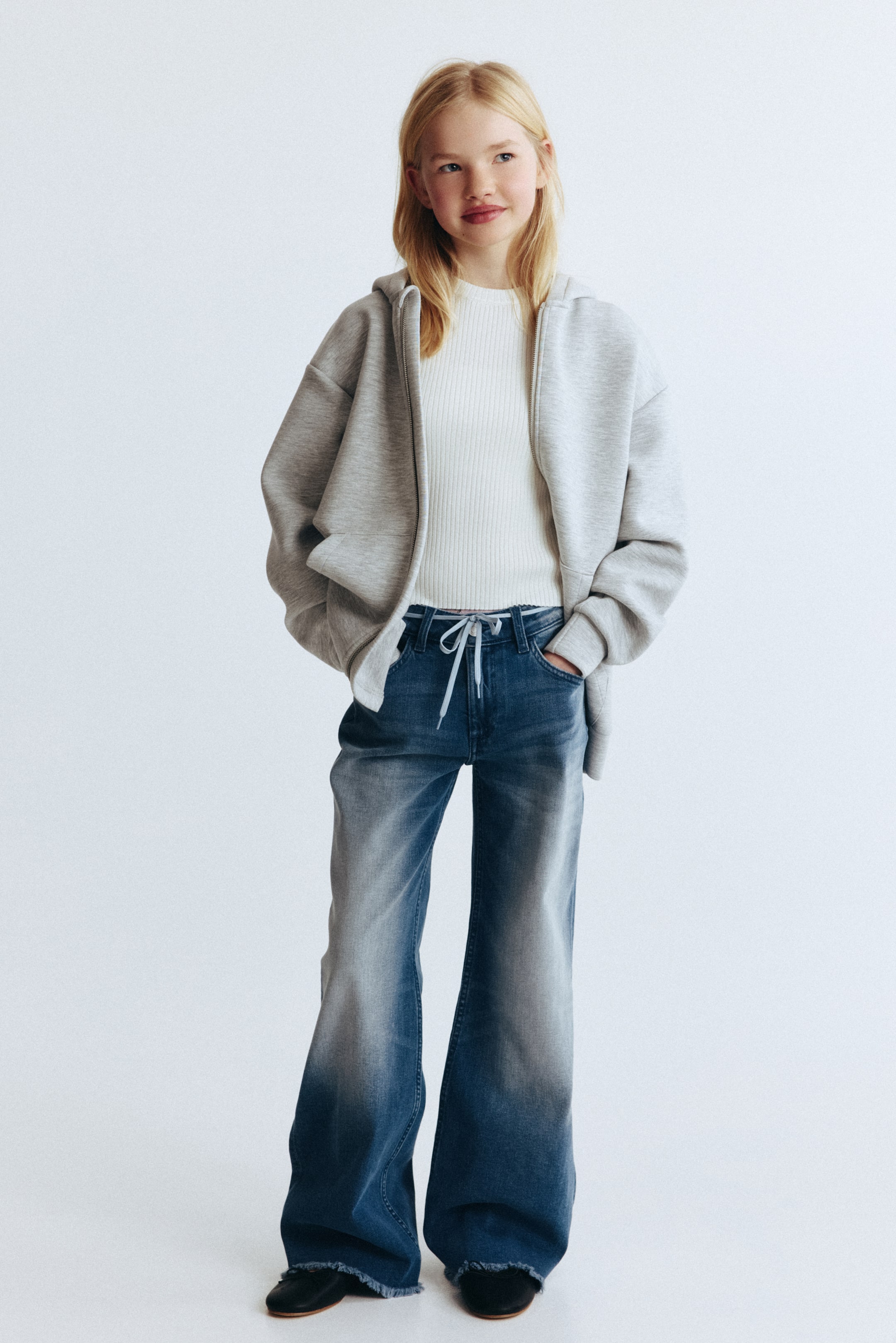 View larger image: A light gray oversized zippered hoodie is presented open over a white ribbed top. Blue wide-leg jeans are worn, featuring a faded wash, a visible drawstring waist, and frayed hems.