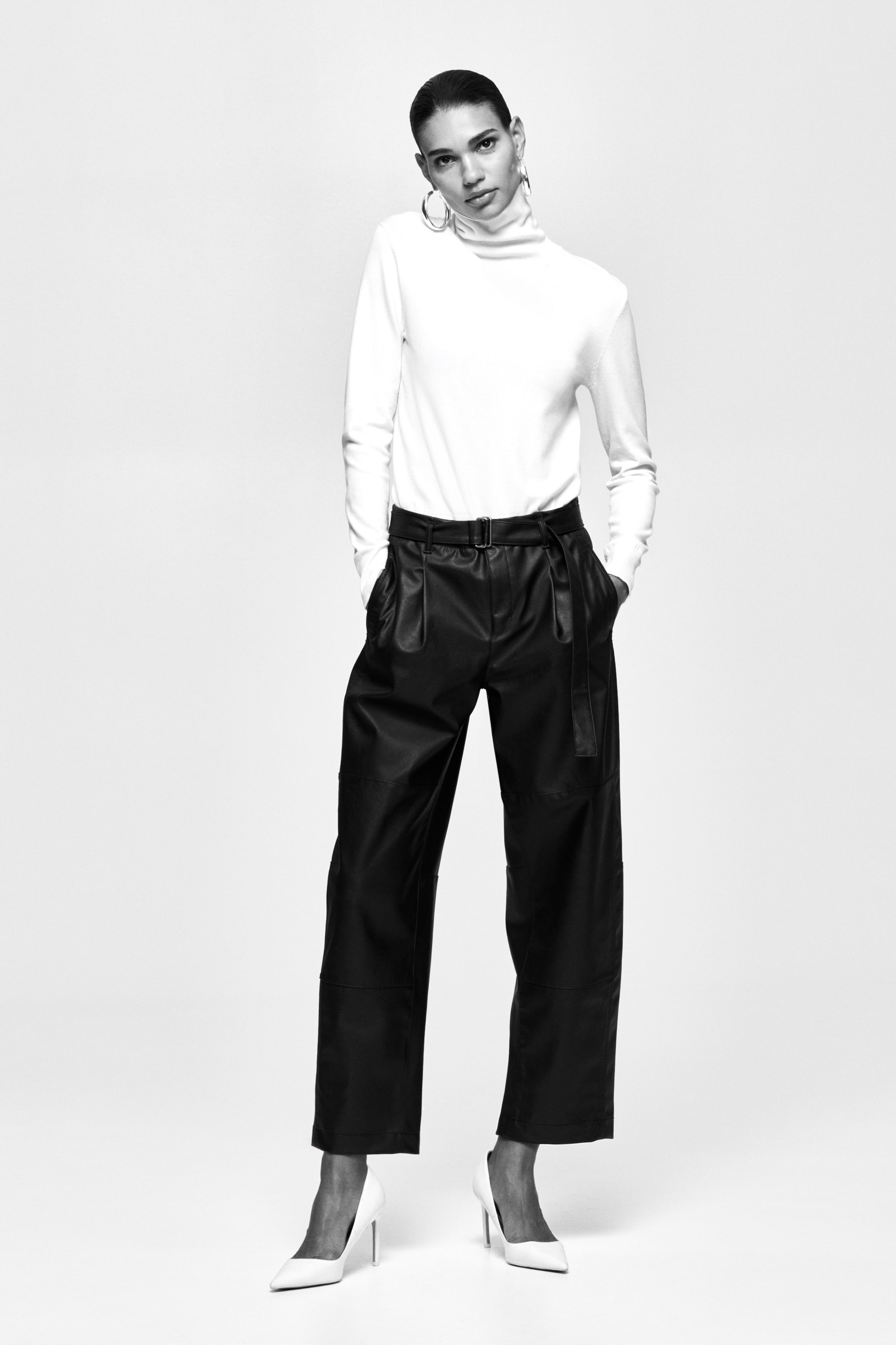 View larger image: Black leather trousers, showcasing pleats at the waist, a self-fabric belted waist, and a wide, cropped leg, are styled by the model with a white turtleneck and white pointed-toe heels.