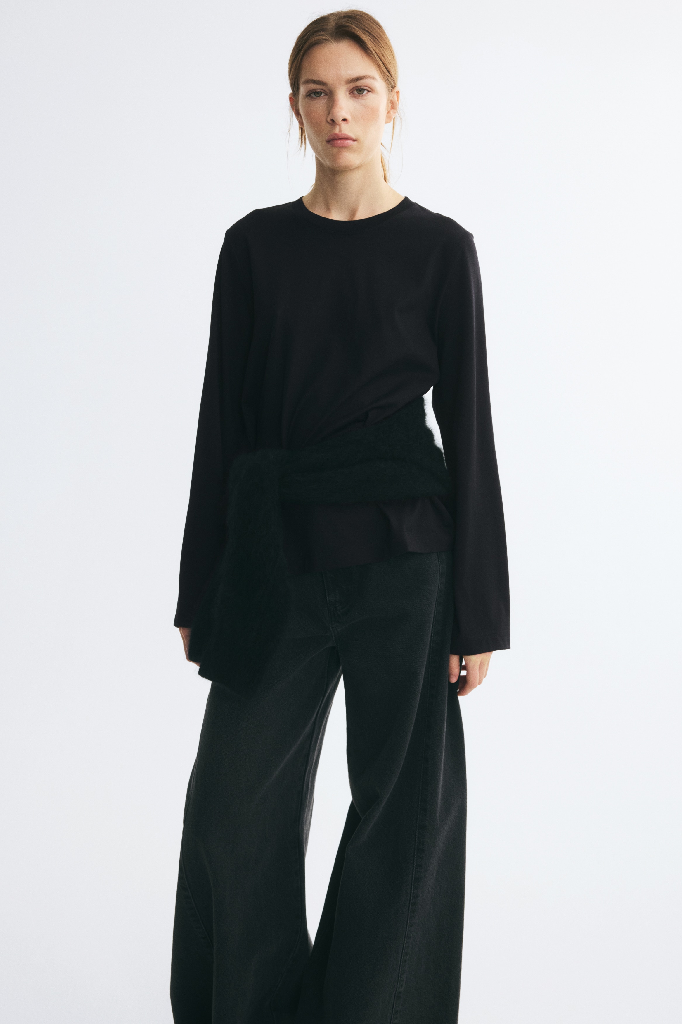 Um top de jersey de algodÃ£o preto estÃ¡ pendurado de frente, apresentando um decote redondo com acabamento em rib e mangas compridas de corte amplo com bainhas direitas. A peÃ§a exibe uma silhueta de corte regular, com uma bainha inferior de acabamento limpo.