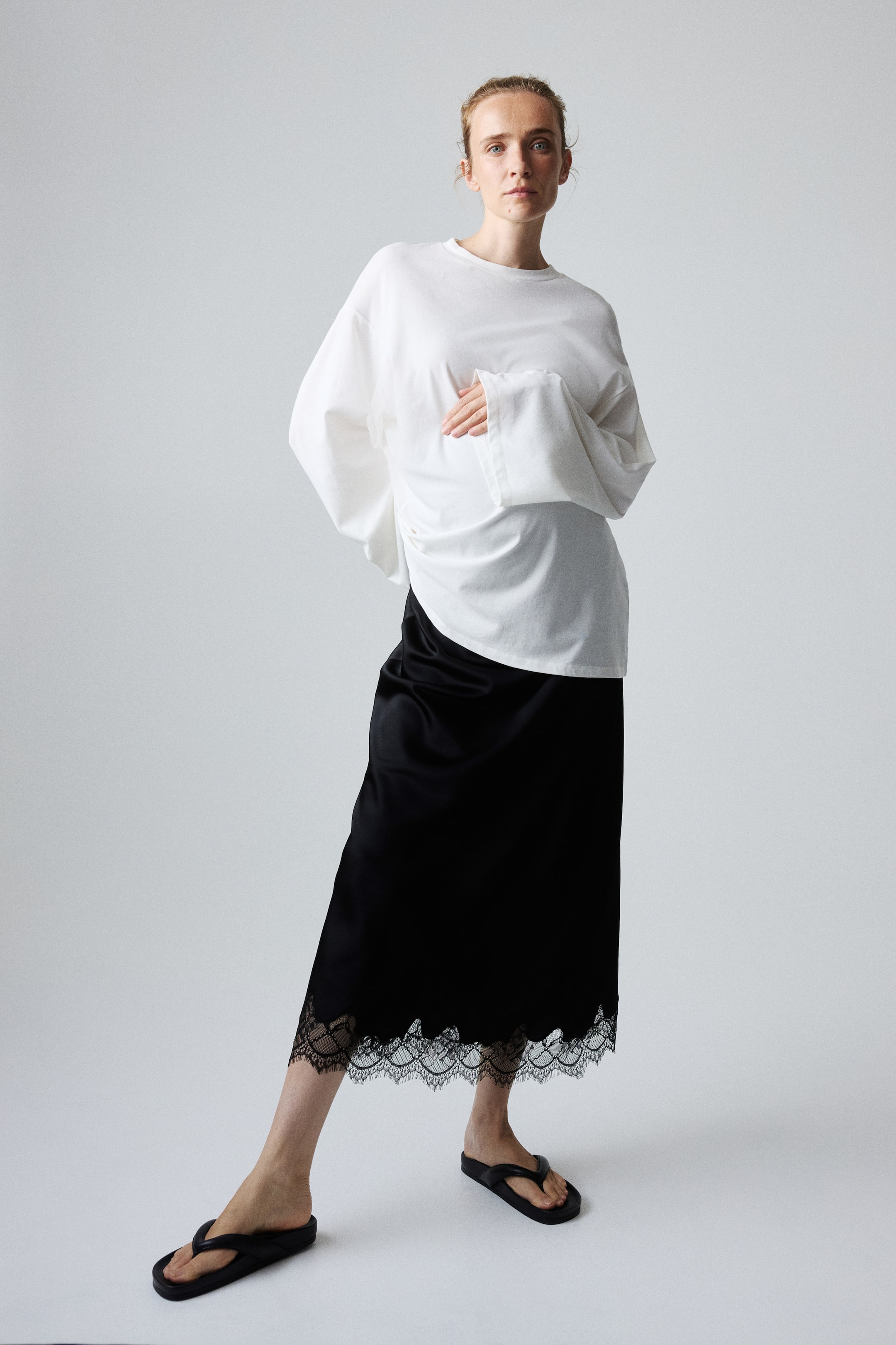 View larger image: A calf-length black satin skirt, featuring a softly draping silhouette and a scalloped, lace-trimmed hem, is being worn by the model. A white oversized top is layered over the skirt’s upper portion.