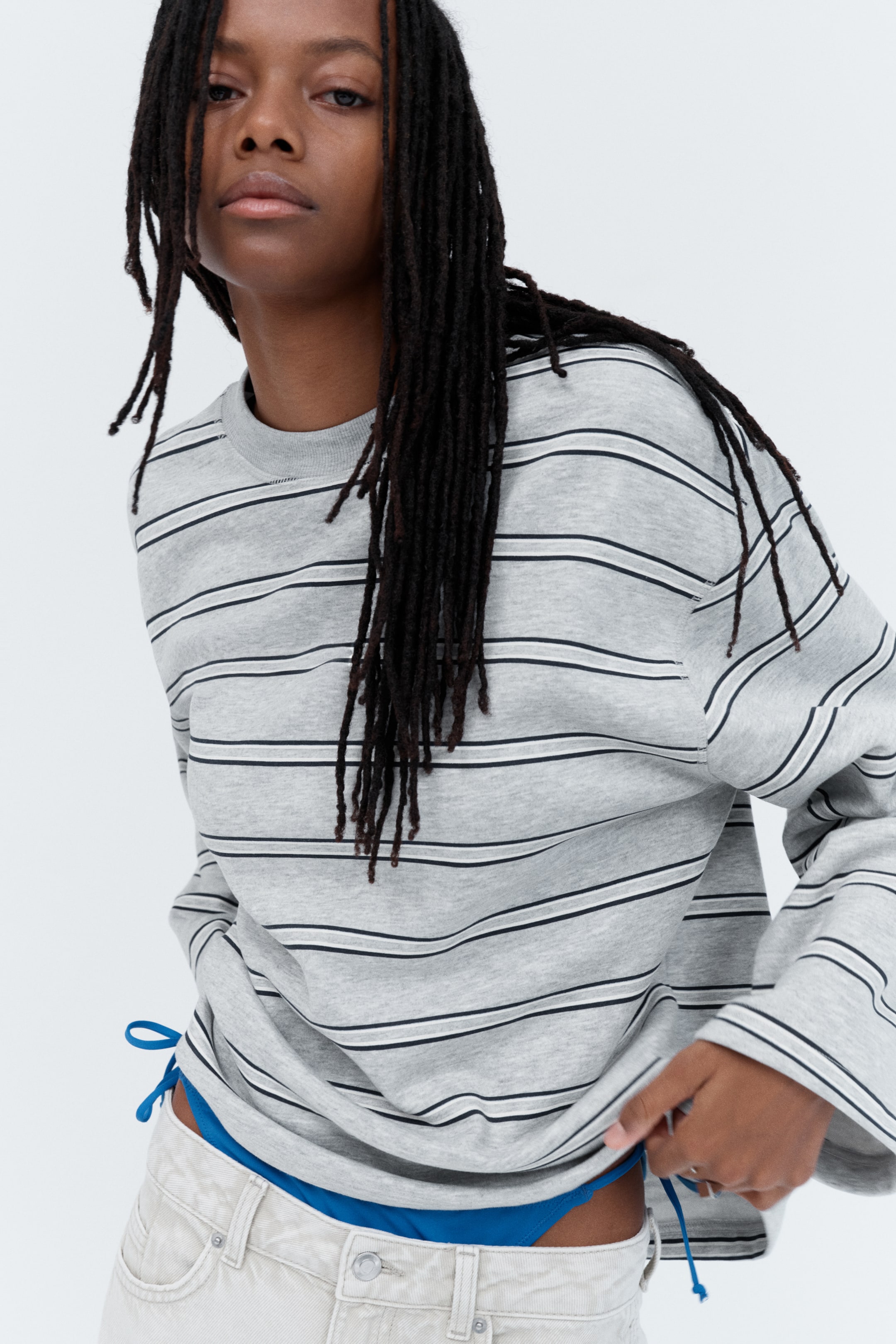 Größeres Bild anzeigen: Das Model trägt einen oversized Sweatshirt mit langen Ärmeln in Grau mit dünnen schwarzen horizontalen Streifen. Das Model hält den Saum des Sweatshirts, das überfallene Schultern und einen gerippten Halsausschnitt aufweist, über einer hellen Hose und einem blauen Unterbekleidungsstück.