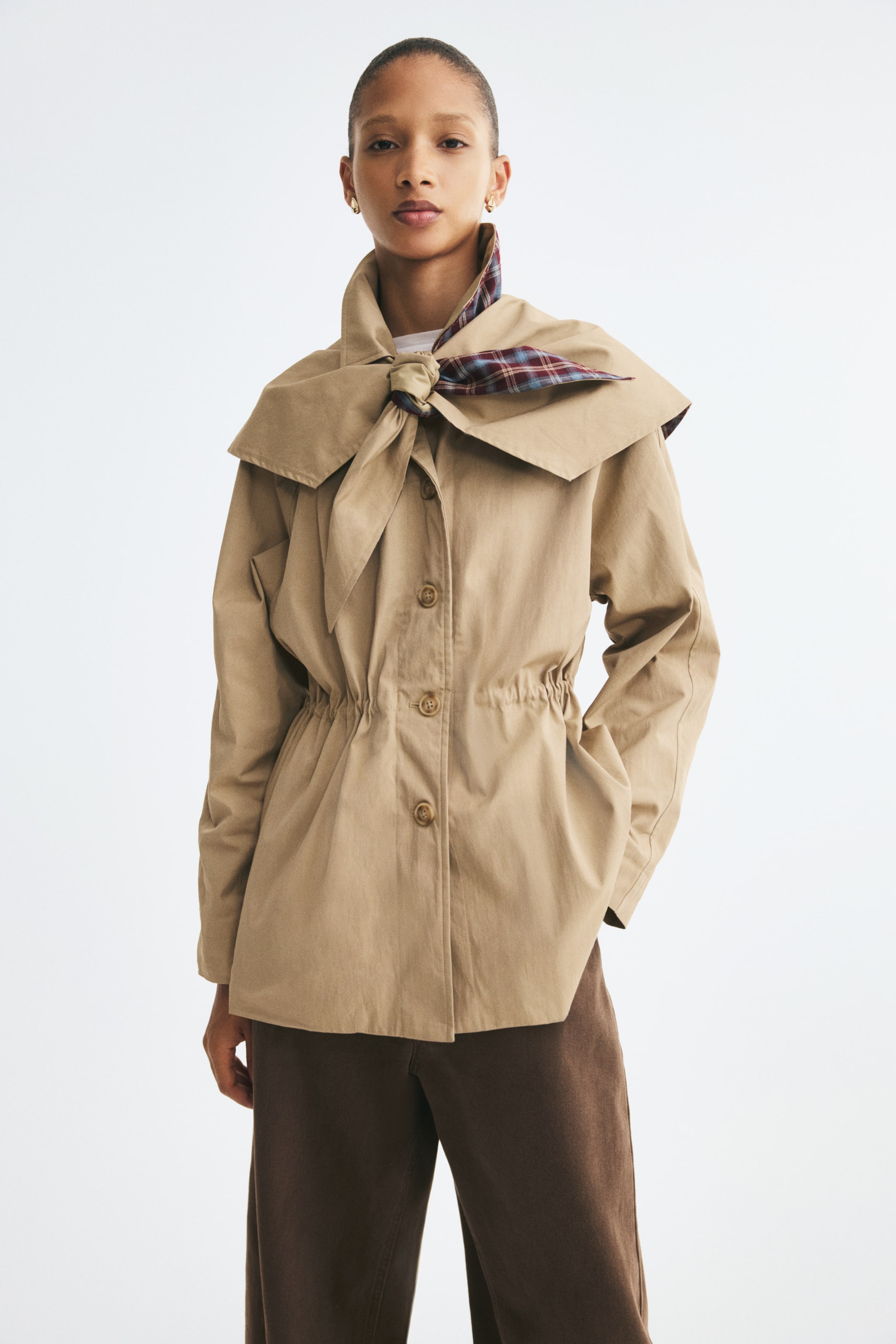 Visualizza immagine più grande: Una giacca in twill beige è mostrata frontalmente, con chiusura con bottoni, maniche lunghe e vita regolabile con effetto arricciato a coulisse. La giacca è abbinata a una sciarpa triangolare staccabile coordinata, annodata al colletto, che mostra una fodera a quadri.