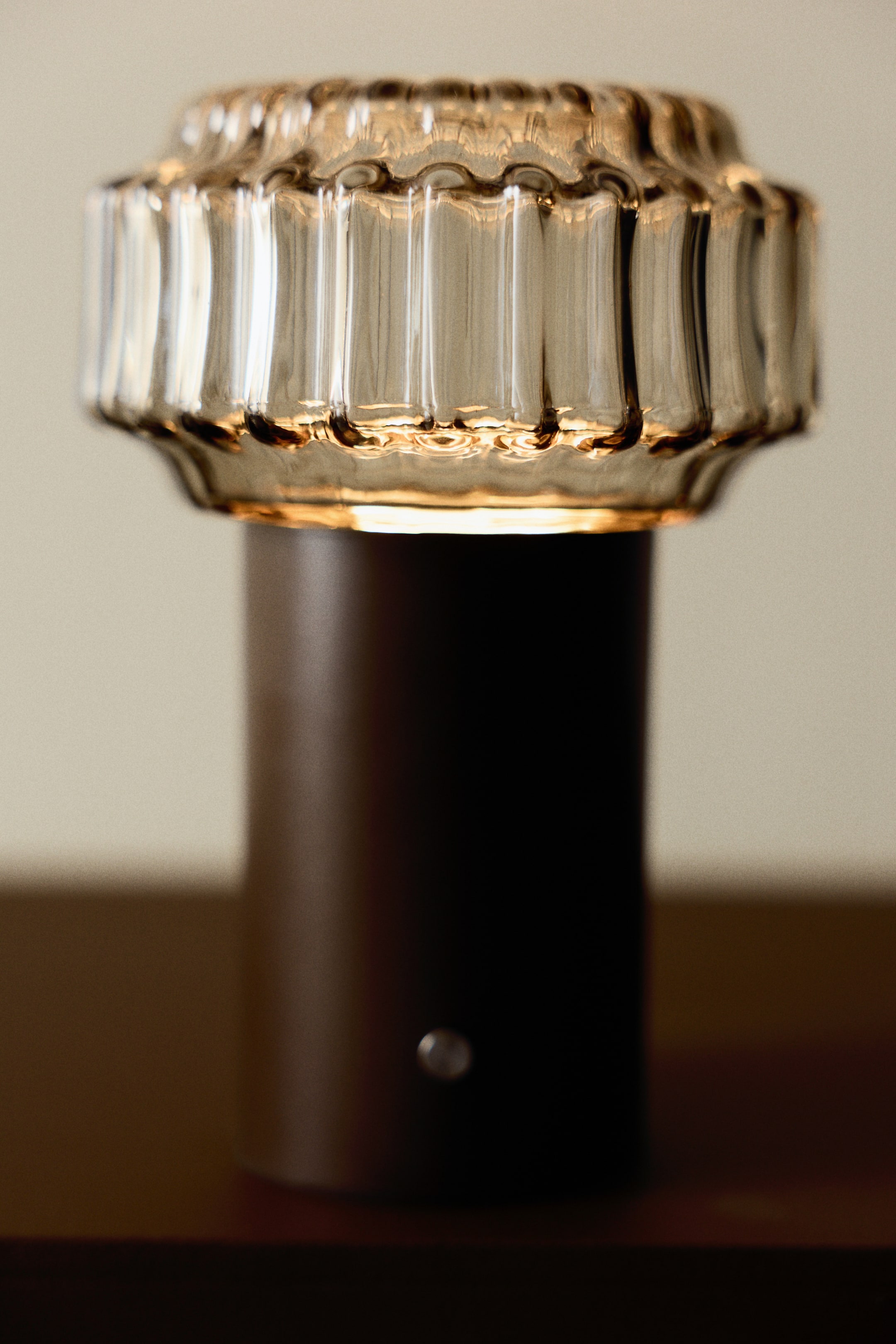 Agrandir l'image: Une lampe de table est dotée d'un abat-jour en verre ambré cannelé qui brille intensément, reposant sur une base cylindrique brun foncé. Un petit bouton argenté est visible à l'avant de la base.
