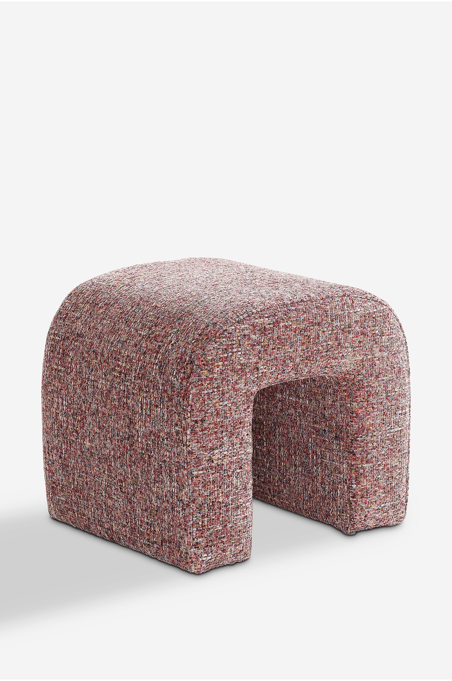 Un pouf a forma di U rivestito in un tessuto intrecciato materico, multicolore, nei toni del rosso e del rosa, Ã¨ mostrato da unâangolazione a tre quarti. Due riviste e una piccola ciotola rotonda in pietra sono appoggiate sulla sua superficie superiore curva.