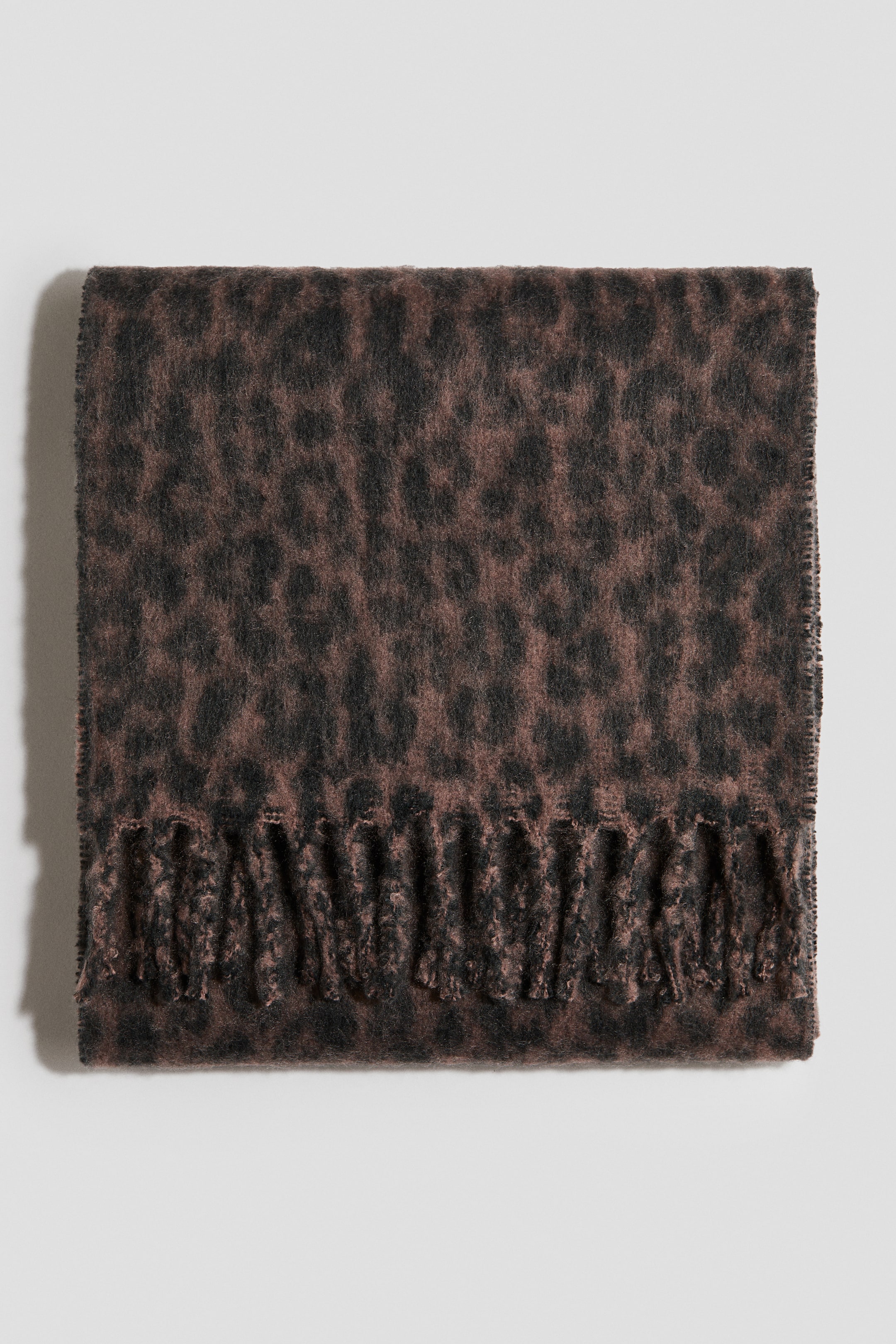 Ver imagen más grande: Una bufanda suave y esponjosa presenta una base marrón apagada con manchas de estampado de leopardo negras. Está doblada en plano, mostrando los extremos con flecos a lo largo del borde inferior.