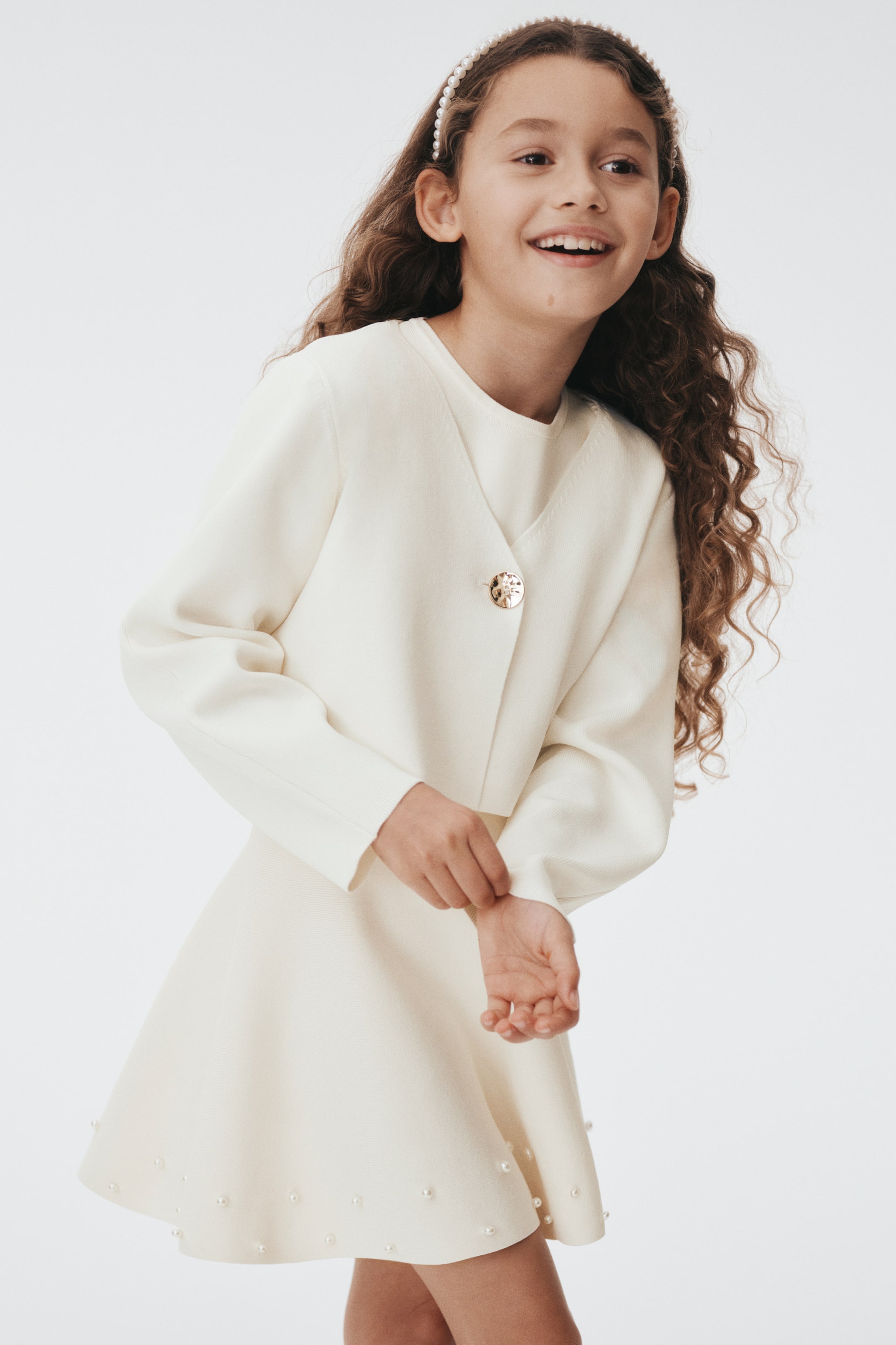 Visa större bild: Ett krämfärgat, tvådelat set presenteras från en framifrån och höger vinkel. Den korta jackan har en V-ringning, långa ärmar med ribbade muddar och en enda, guldtonad, texturerad knapp vid framstängningen. Den matchande A-linjeformade kjolen är dekorerad med små, runda pärlliknande pärlor längs dess fåll.