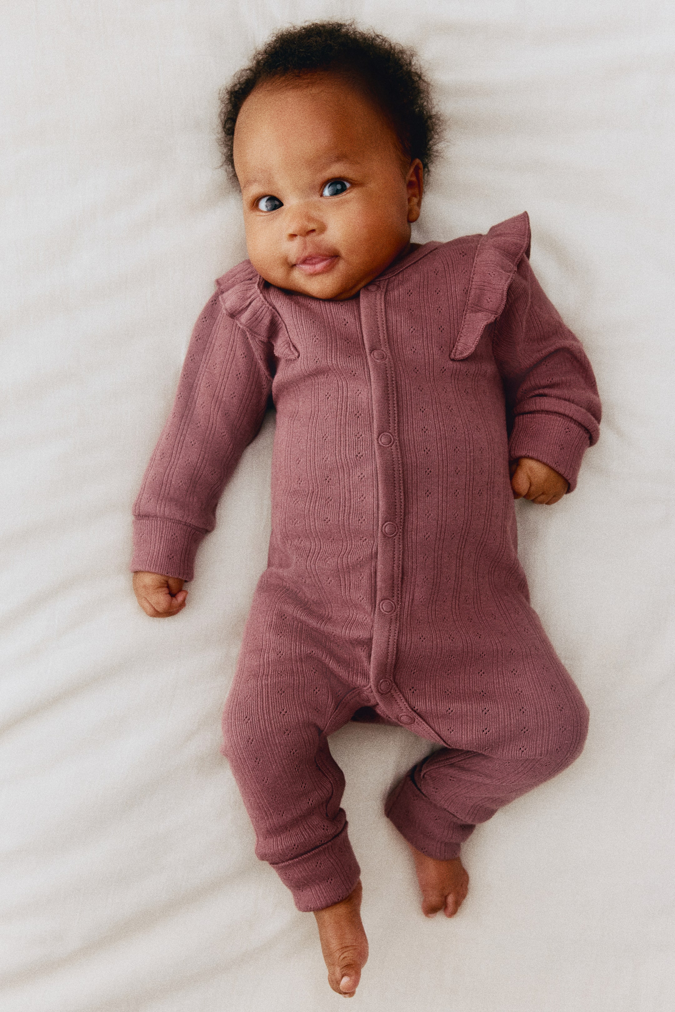 Agrandir l'image: Une combinaison pour bébé en maille pointelle rose poudré est présentée de face, avec des volants aux épaules et une fermeture par boutons-pression לאורך du centre. La combinaison est conçue avec des manches longues et des jambes se terminant par de très longs poignets côtelés repliés.