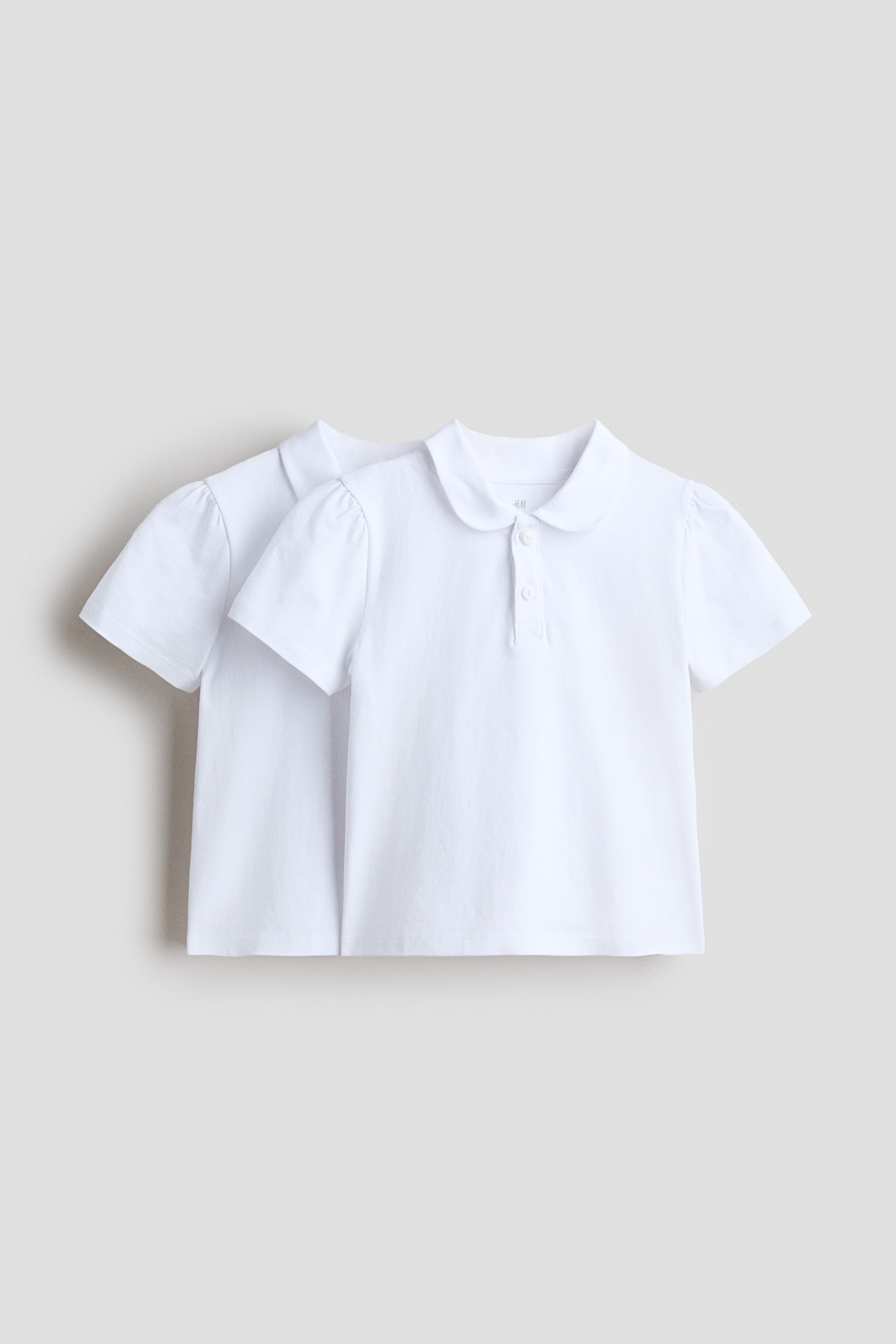 View larger image: Two white polo shirts are displayed, slightly overlapping. Each shirt features a rounded Peter Pan collar and a three-button placket. They also have short, slightly puffed sleeves.