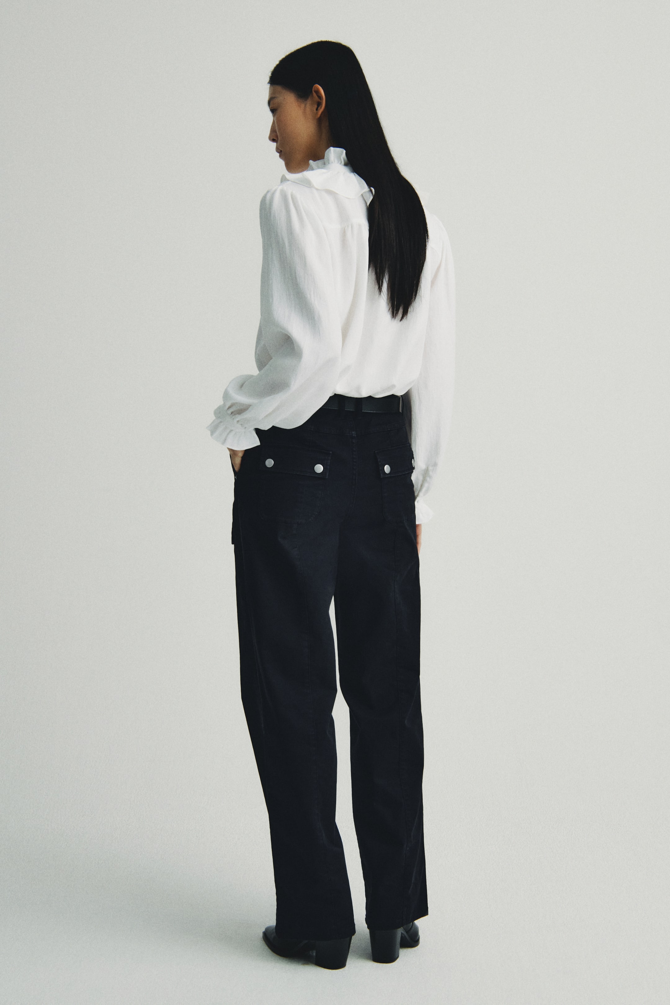 Xem ảnh lớn hơn: Một chiếc áo blouse trắng, với cổ đứng bèo nhún và cổ tay áo bèo nhún, đang được giới thiệu từ góc nhìn phía sau bên trái. Nó được sơ vin vào quần ống rộng nhung tăm màu đen, được thiết kế với hai túi có nắp, mỗi cái được cố định bằng một nút màu bạc phía sau, và đang được kết hợp với một chiếc thắt lưng màu đen.