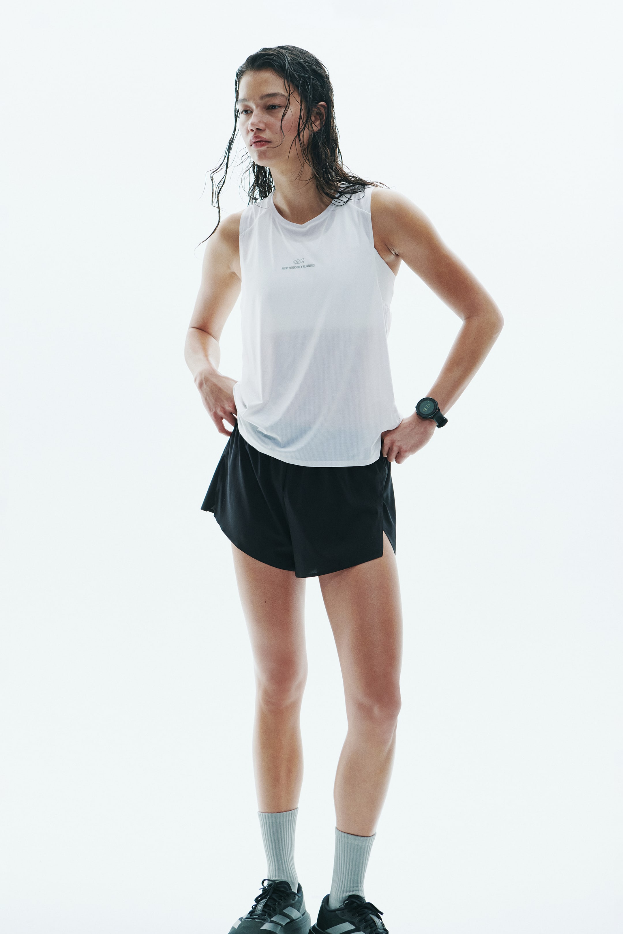 Ampliar la imagen: Una playera deportiva blanca sin mangas, con cuello redondo y un pequeño gráfico en el pecho, es usada por la modelo junto con shorts negros para correr con aberturas laterales. La modelo posa con las manos apoyadas en las caderas, complementando la ropa deportiva con un reloj negro, calcetas grises tipo crew y tenis negros para correr.