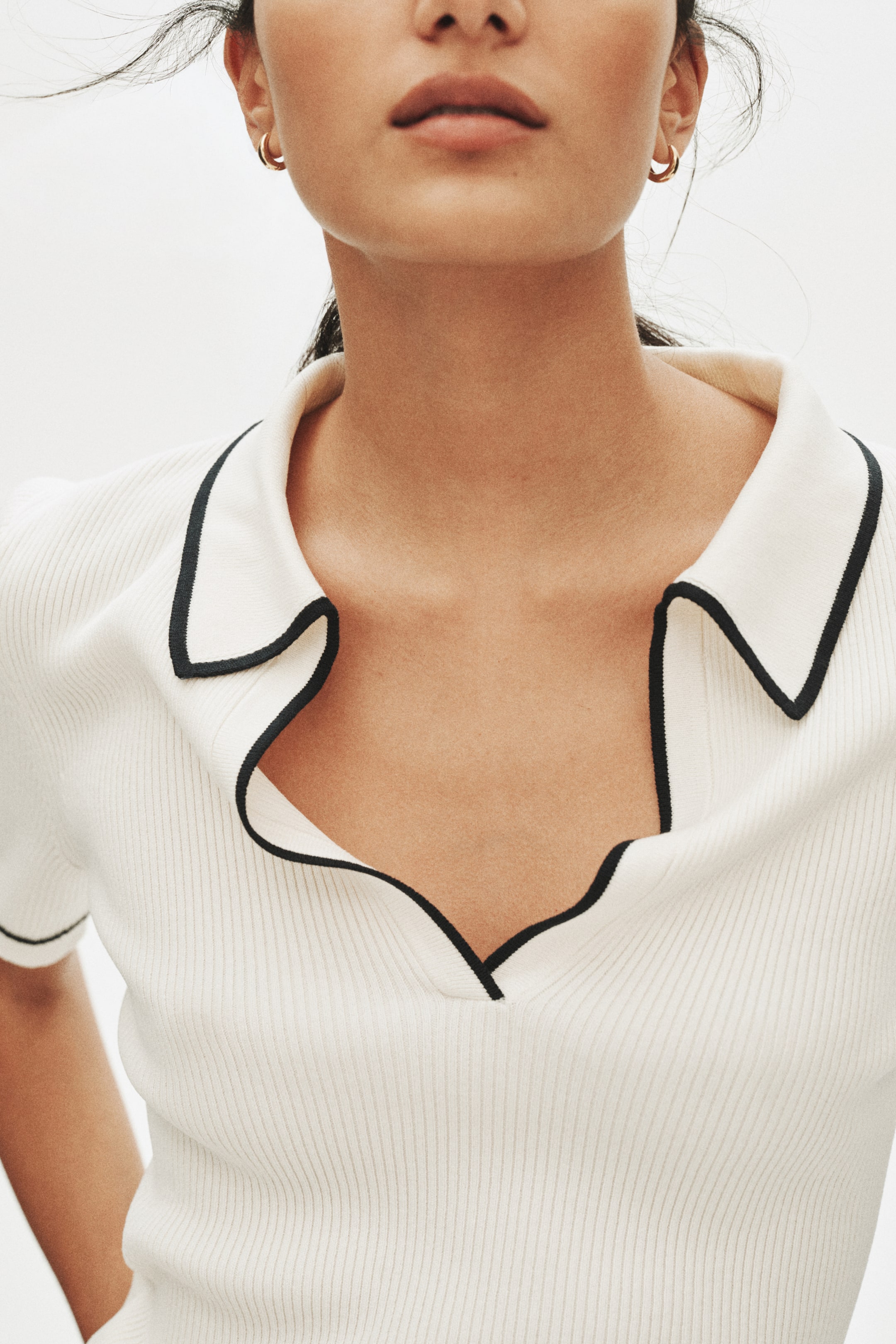 View larger image: A cream-coloured ribbed-knit top features an open collar with black contrast piping. Its short sleeves are also detailed with black piping along the cuffs.