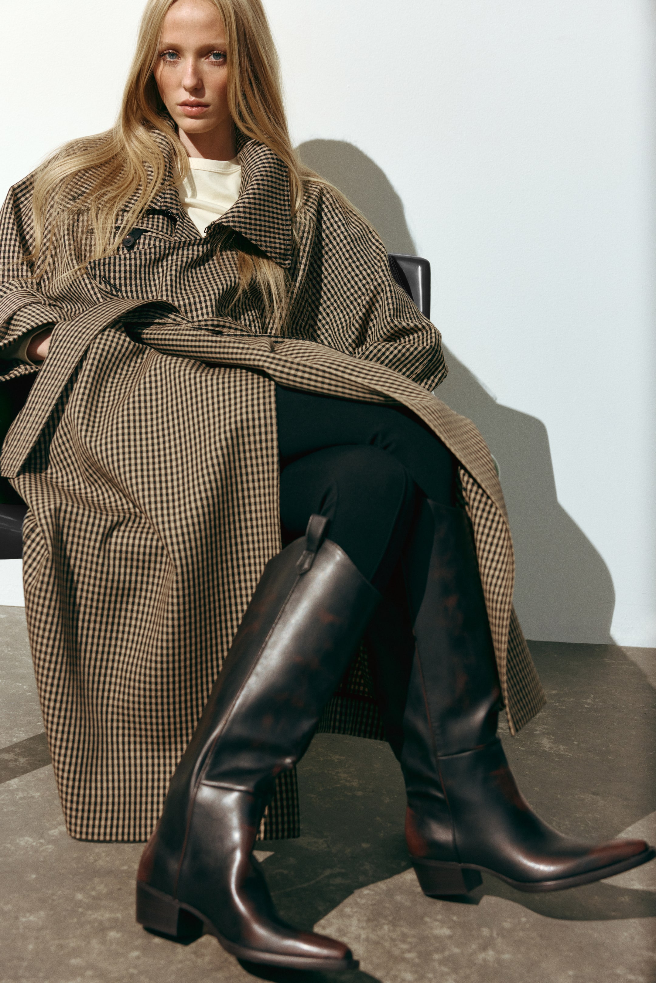 Größeres Bild ansehen: Dunkelbraune kniehohe Lederstiefel mit einer polierten Oberfläche und niedrigen Blockabsätzen werden von dem Model getragen. Die Stiefel sind mit schwarzen Leggings und einem langen, karierten braunen und schwarzen Trenchcoat kombiniert.