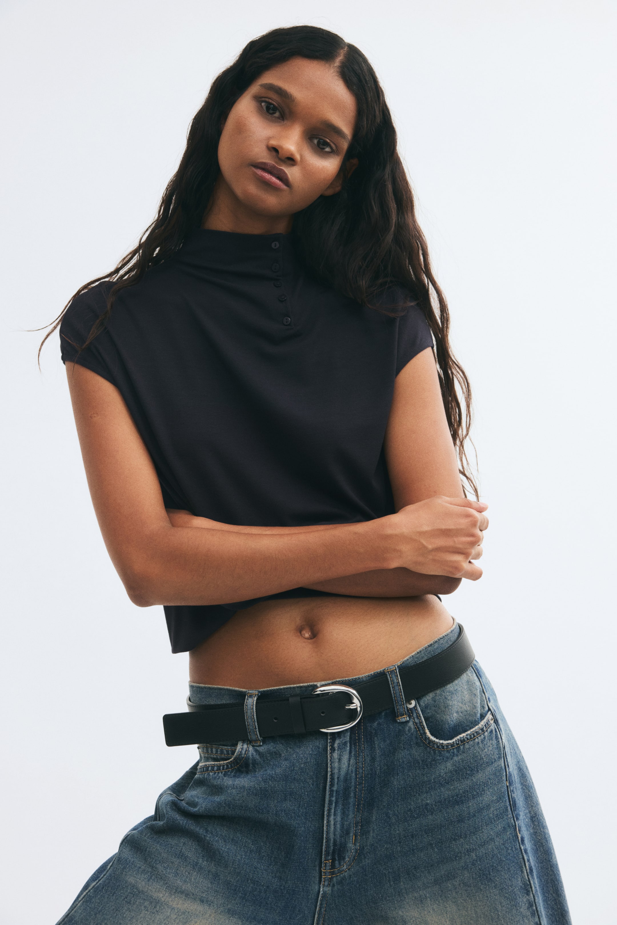 View larger image: The model is wearing a dark blue cropped top featuring a high neck with button details on one side and short cap sleeves. The top is styled with blue jeans and a black belt, revealing the midriff.