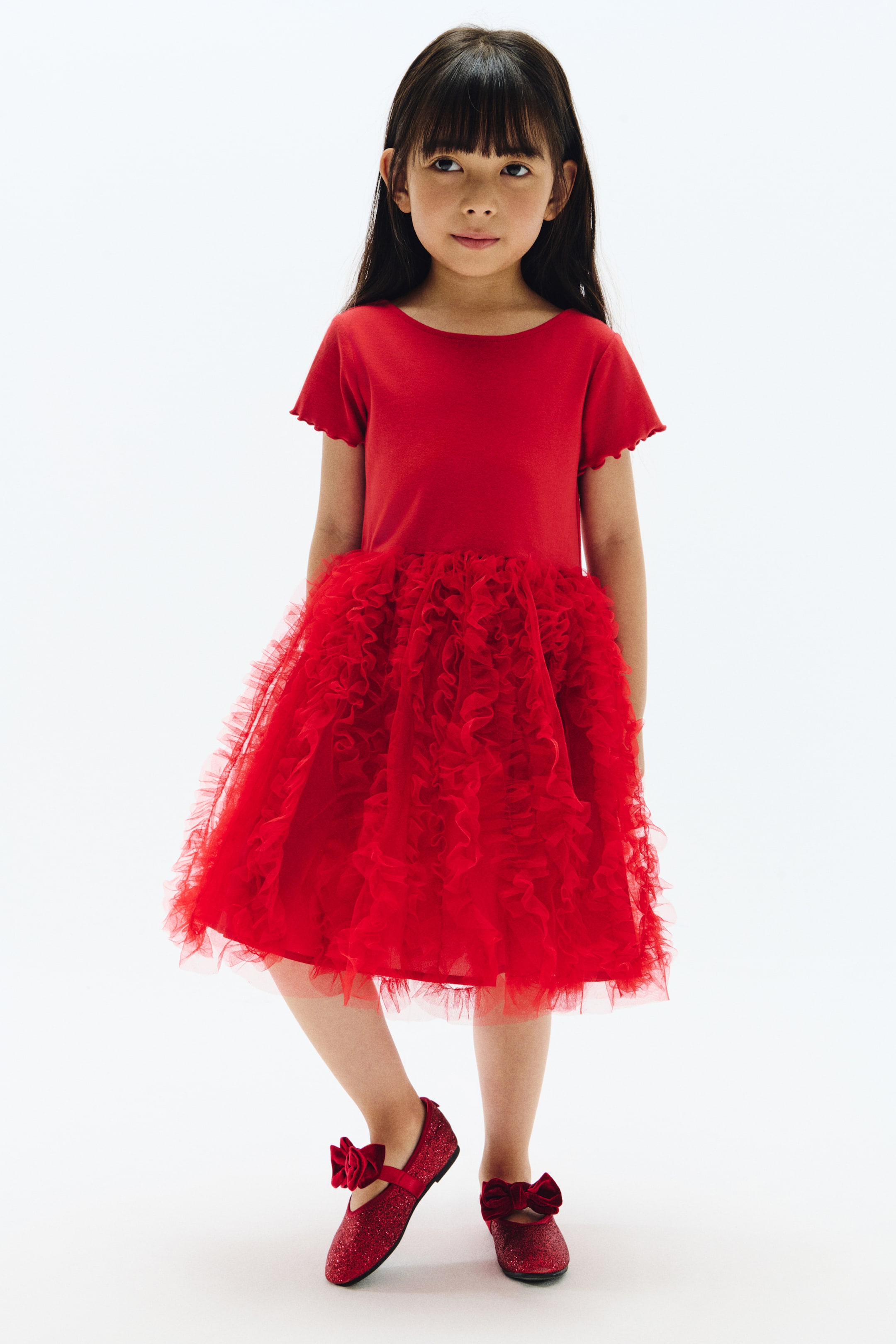 View larger image: A red dress featuring a solid bodice with scalloped short sleeves and a boat neckline, paired with a voluminous ruffled tulle skirt. The look is completed with red glitter Mary Jane flats, each finished with a red bow.
