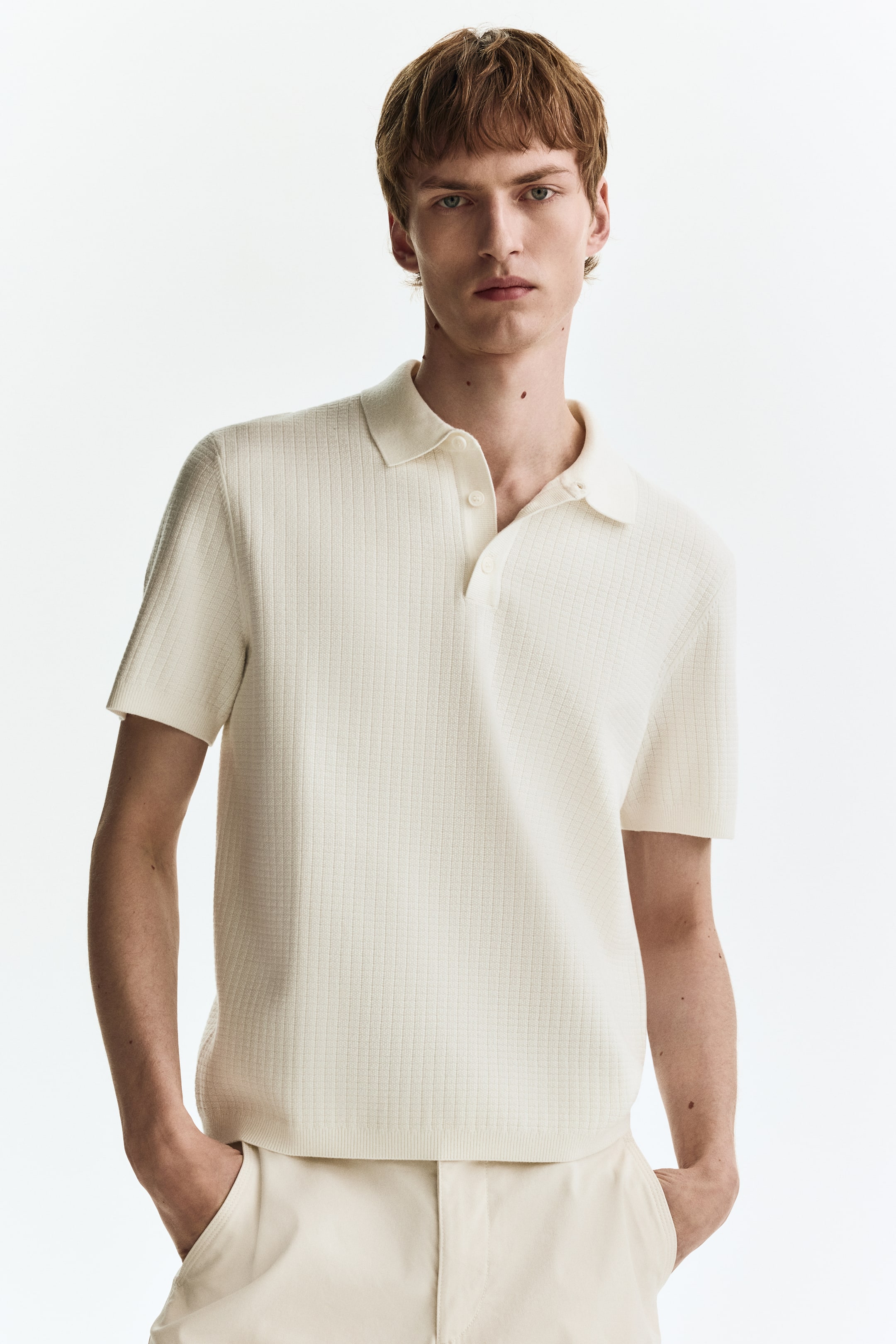 View larger image: An off-white knitted polo shirt is facing forward, featuring a classic collar, a two-button placket, and short sleeves with finished cuffs. The shirt showcases a textured, square-grid pattern throughout its fabric and a finished bottom hem.