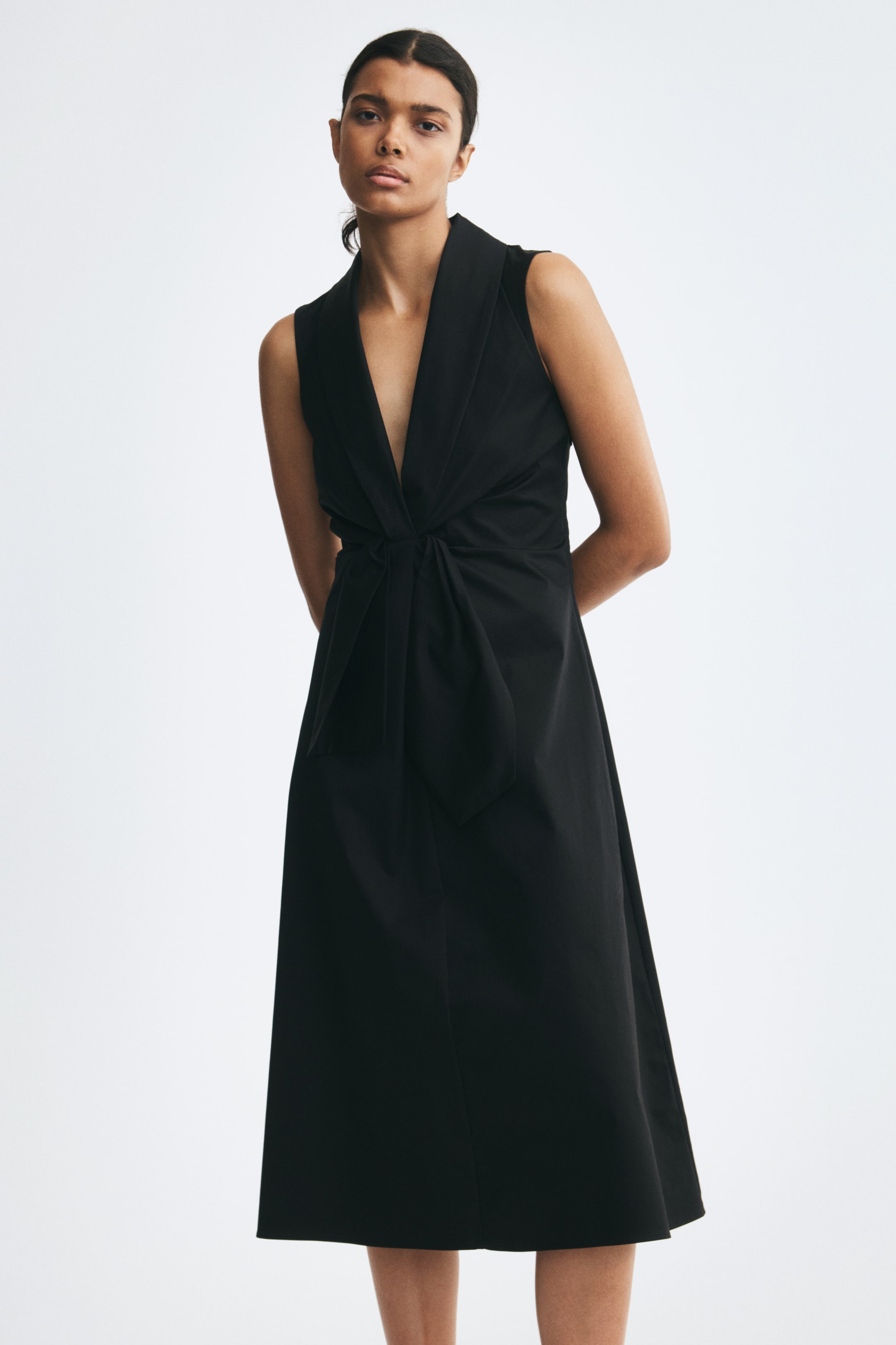 View larger image: The model is wearing a black, calf-length, sleeveless dress. The dress features a deep V-neck with wide lapels, a fitted bodice, and a prominent pleated tie detail at the waist that drapes over a gently flared skirt.