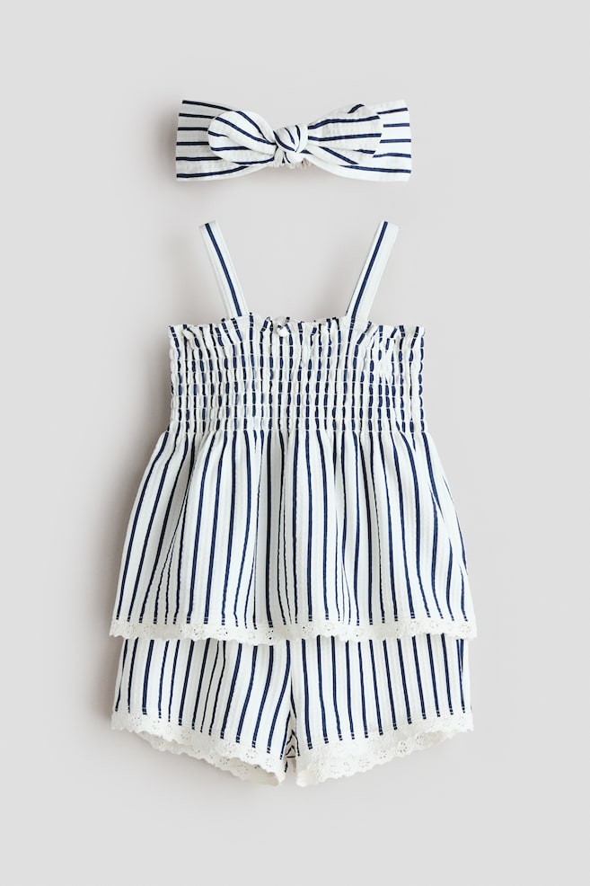 A baby's two-piece outfit and matching headband are displayed from the front. The sleeveless top and shorts feature white fabric with navy vertical stripes and white lace trim at the hems, with the top also showcasing a smocked bodice and thin shoulder straps. The coordinating striped headband ties in a knot. - 1