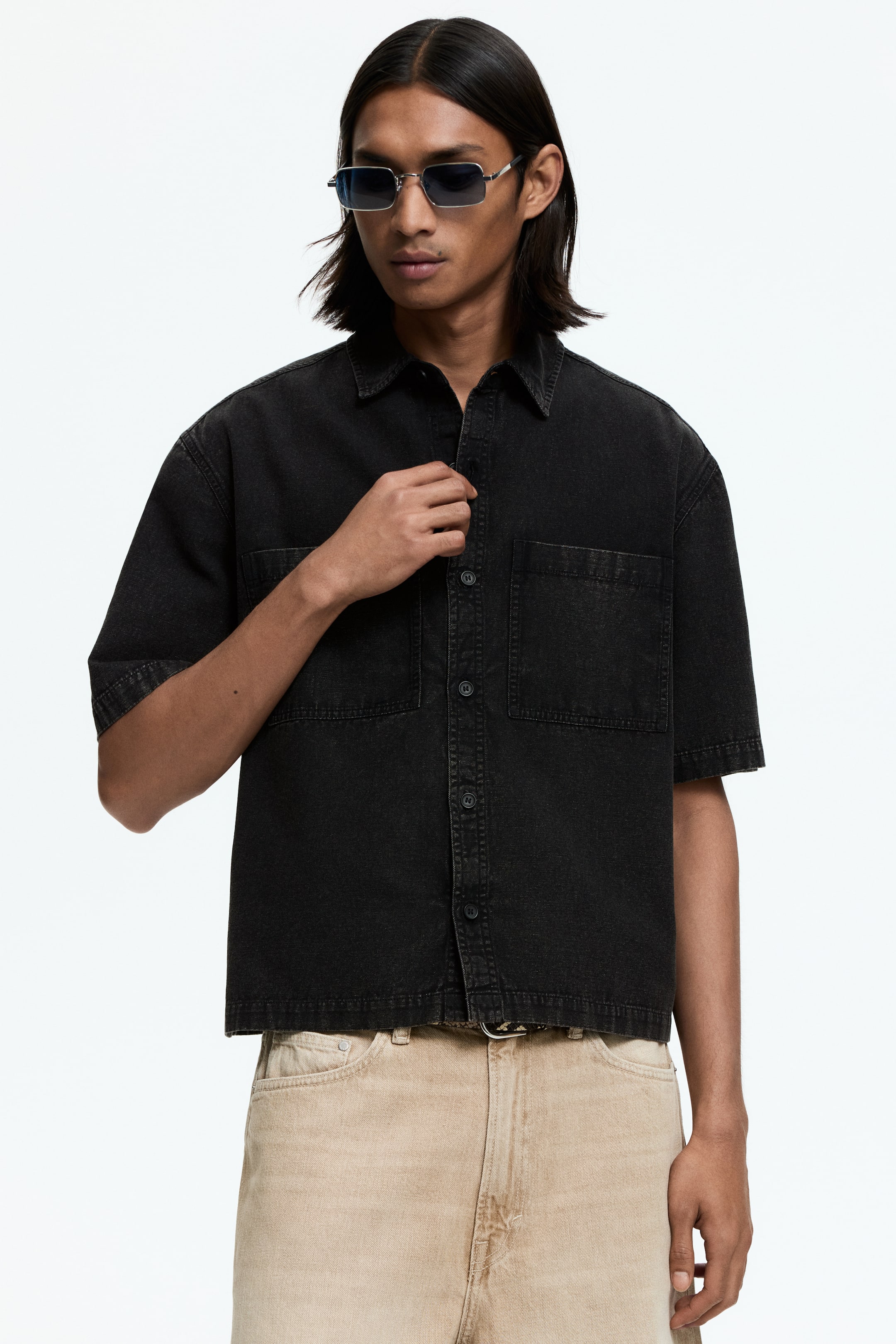 View larger image: The black short-sleeved denim shirt showcases a pointed collar, button-front closure, and two forward-facing patch chest pockets. It is crafted with a relaxed fit and features a cropped, raw hem, with a hand depicted buttoning its front.