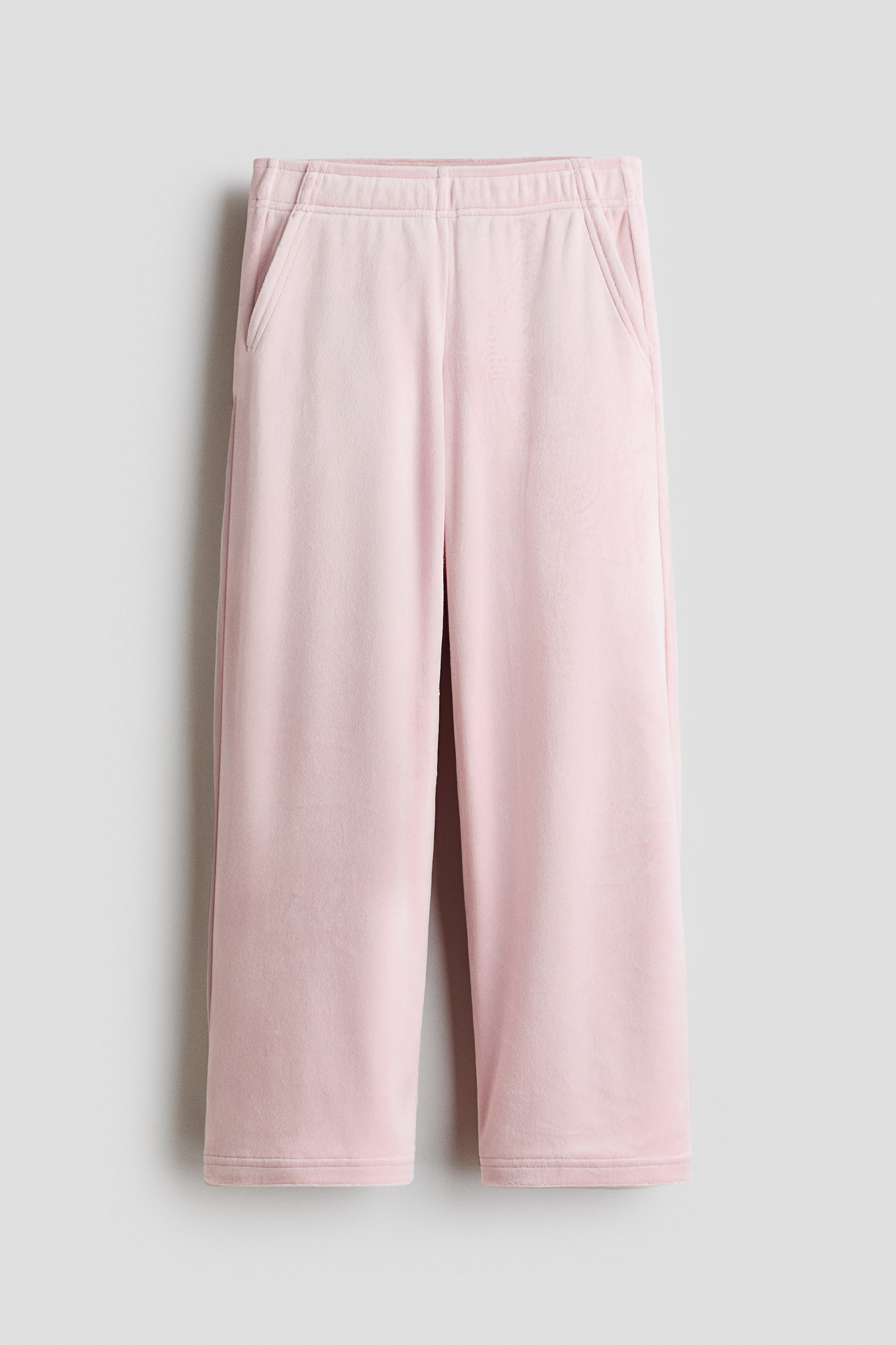 View larger image: A pair of light pink, soft-textured trousers is hanging front-facing, showcasing an elasticised waistband and side slant pockets. The trousers feature a relaxed, wide-leg silhouette that drapes straight down to the hems.