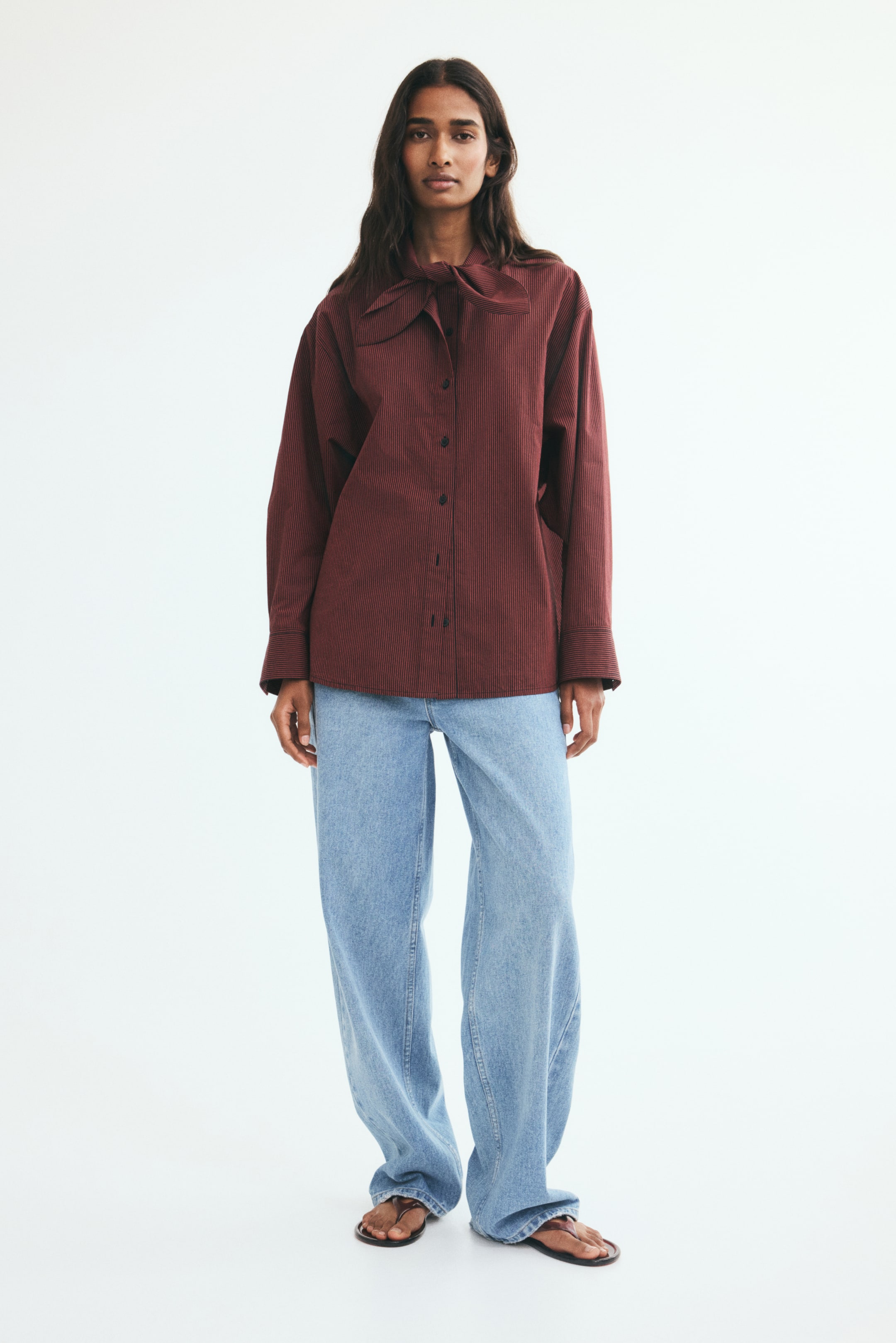 View larger image: The model is wearing an oversized burgundy pinstriped shirt with a prominent bow collar, featuring a button-down front and long sleeves. The straight-cut shirt is styled untucked over light-wash blue denim jeans.