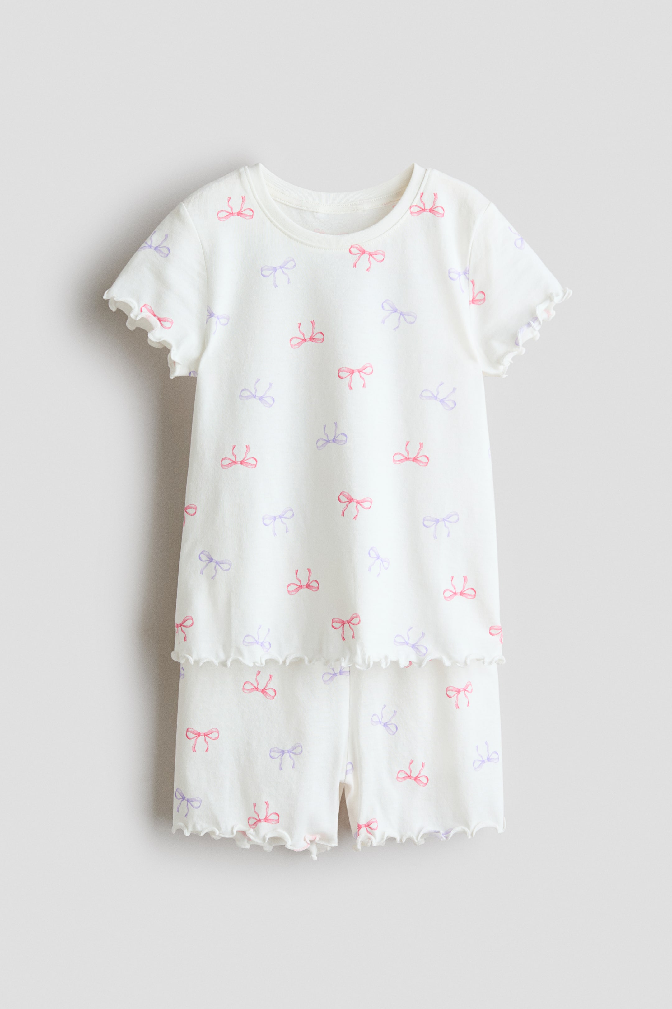 Grösseres Bild ansehen: Ein weisses Pyjama-Set mit einem Muster aus rosa und violetten Schleifen wird von vorne präsentiert. Das kurzärmelige Oberteil verfügt über einen runden, mit Rippbündchen versehenen Halsausschnitt und versäuberte Kanten an den Ärmelabschlüssen und am Saum, während die passenden Shorts versäuberte Kanten an den Säumen haben.