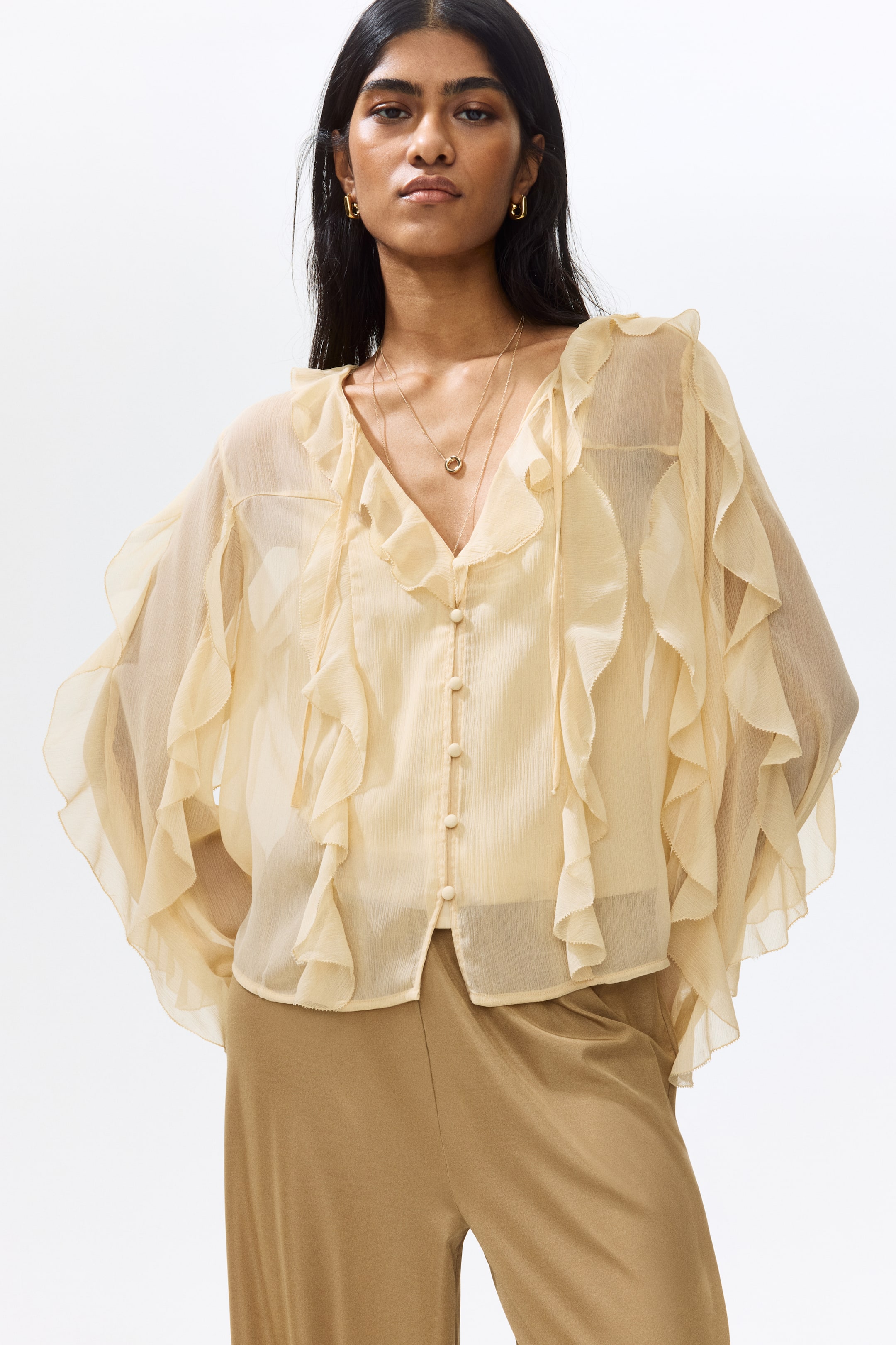View larger image: The model is wearing a cream loose-fit blouse crafted from sheer chiffon, adorned with flounce trims throughout. It showcases a deep V-neckline with slender ties, covered buttons along the front, and long balloon sleeves with elasticated cuffs, styled over tan trousers.