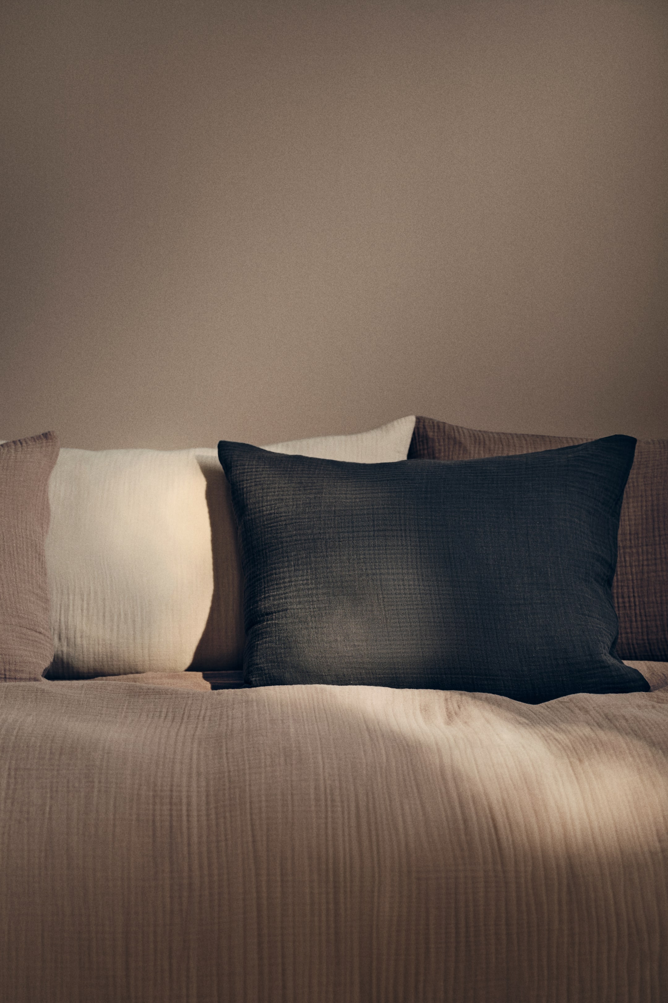 View larger image: "Several rectangular cushions and a coordinating blanket showcase a textured, crinkled fabric. A dark gray cushion is prominently positioned in the foreground, flanked by an off-white cushion and light brown cushions, all facing forward, with the light brown blanket lying beneath them."