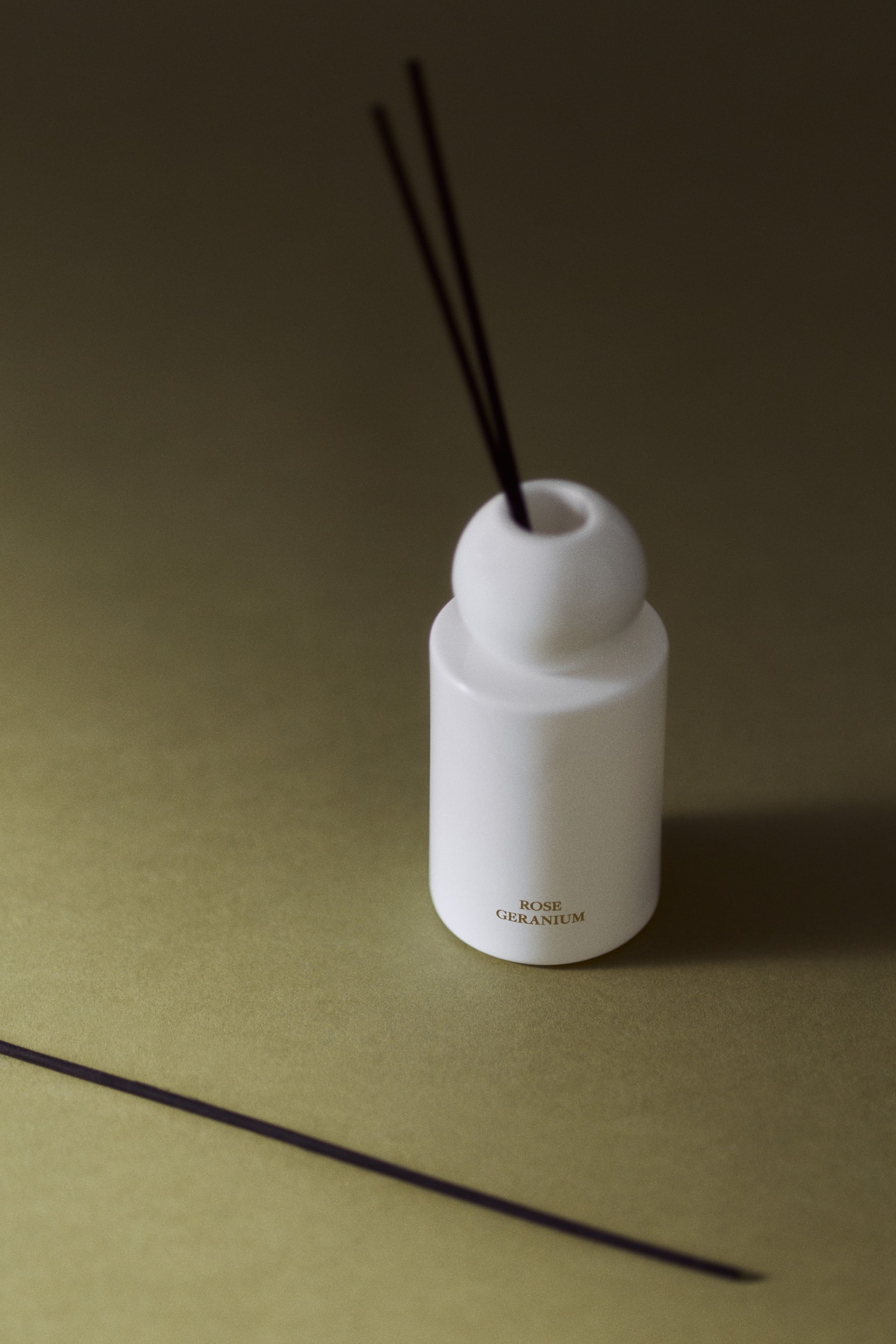 A white cylindrical glass diffuser bottle with a spherical neck is standing upright, with two dark wooden reeds extending from the top opening. The words "ROSE GERANIUM" are printed in gold lettering near the lower front of the bottle. - 1