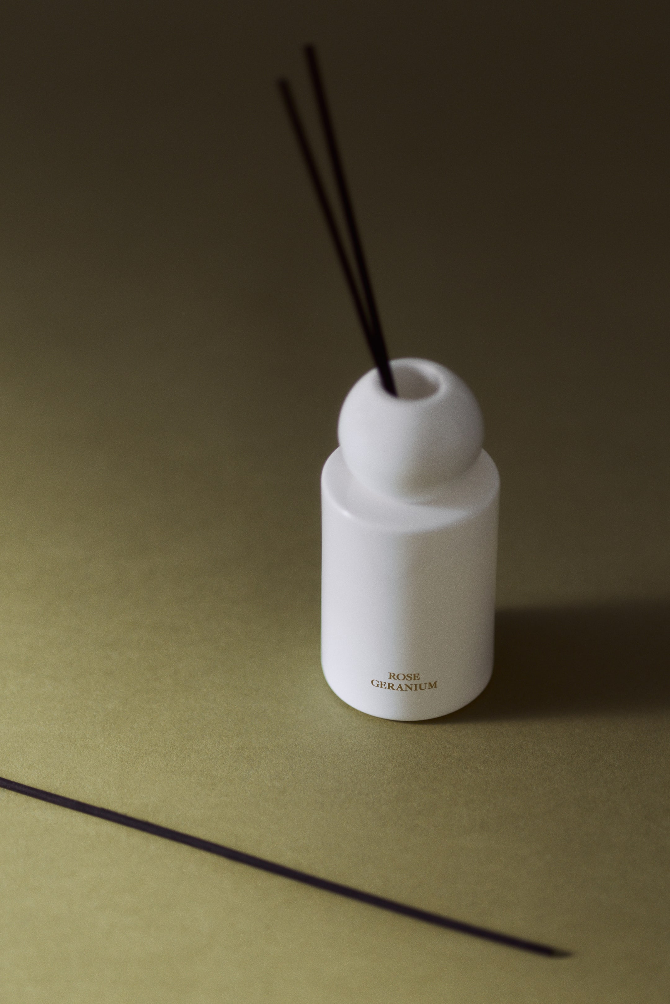 View larger image: A white cylindrical glass diffuser bottle featuring a spherical neck is standing upright, with two dark wooden reeds extending from the top opening. The words "ROSE GERANIUM" are printed in gold lettering near the bottom front of the bottle.