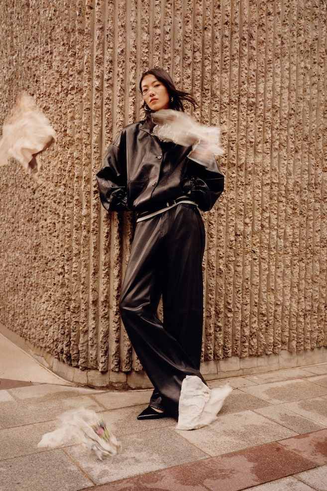 An oversized black jacket in coated fabric is being worn by the model, paired with matching wide-leg trousers. The jacket features a buttoned front, dropped shoulders, and elasticated cuffs and hem. - 1