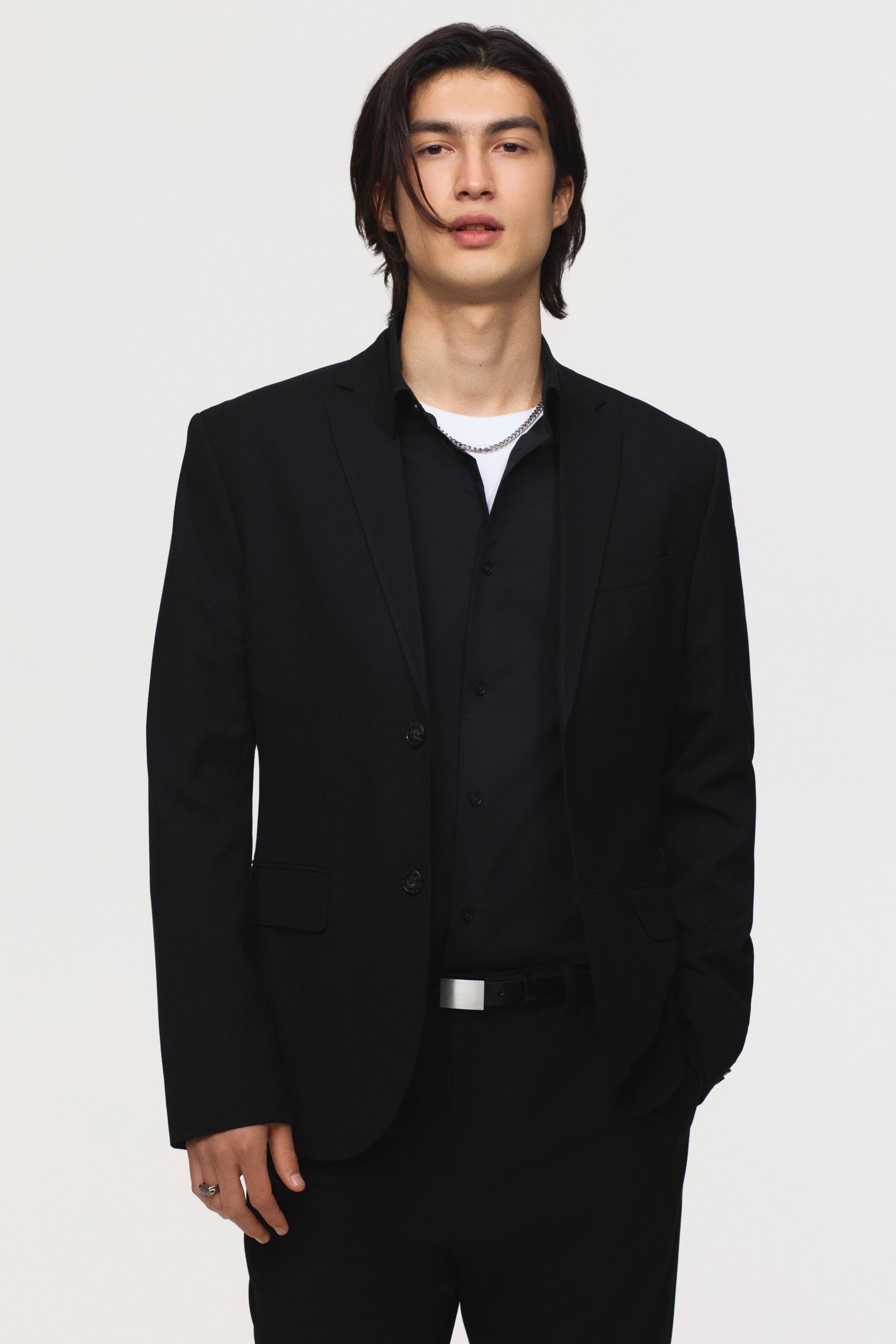 View larger image: A black single-breasted suit jacket features notch lapels, two front buttons, and flap pockets. It is styled over a black collared shirt, a white tee, and a silver chain, paired with matching black trousers and a black belt with a silver buckle.