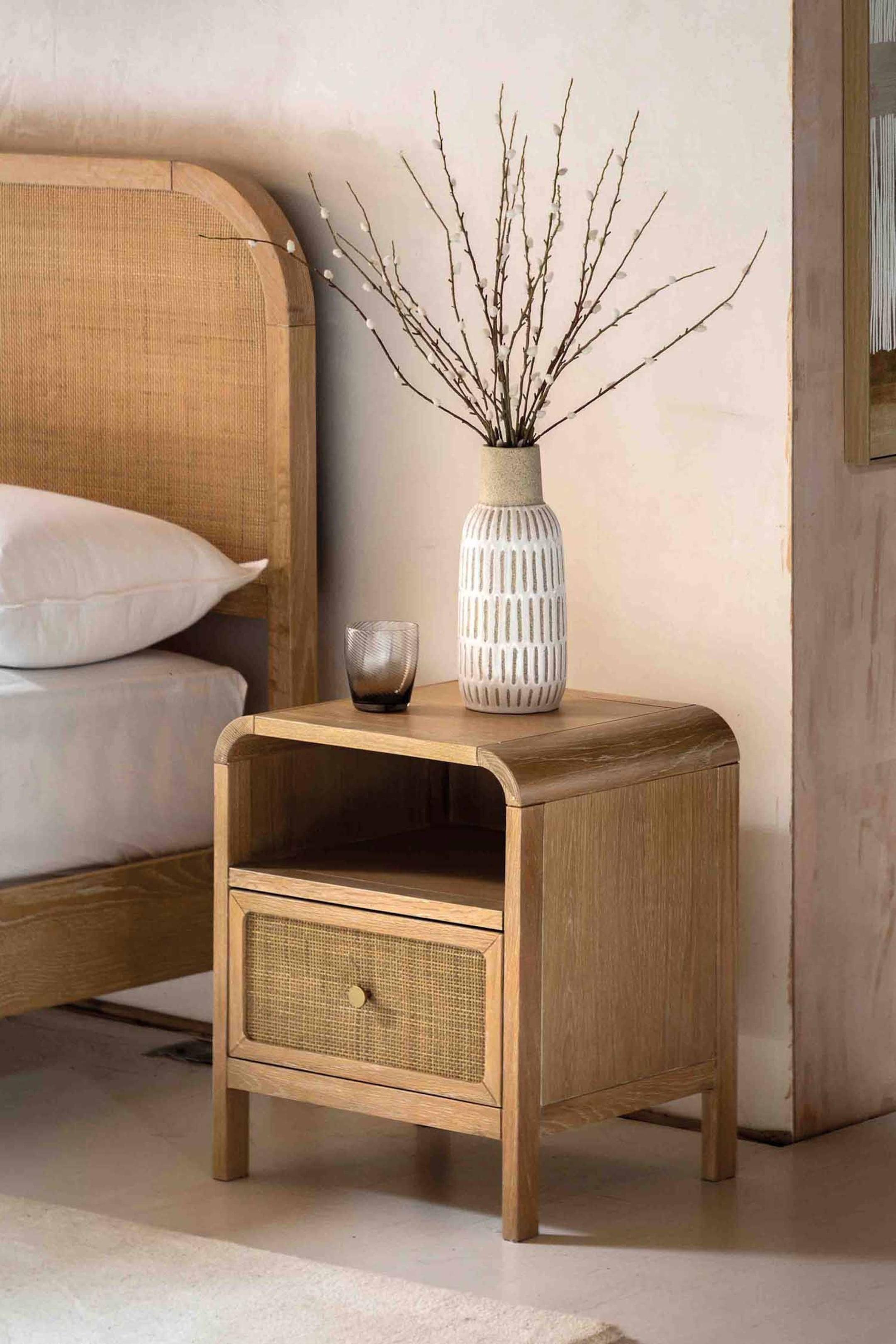 View larger image: A light-toned wooden bedside table with visible grain features a rounded top surface, an open storage shelf, and a lower drawer with a woven cane front and a small, round knob.